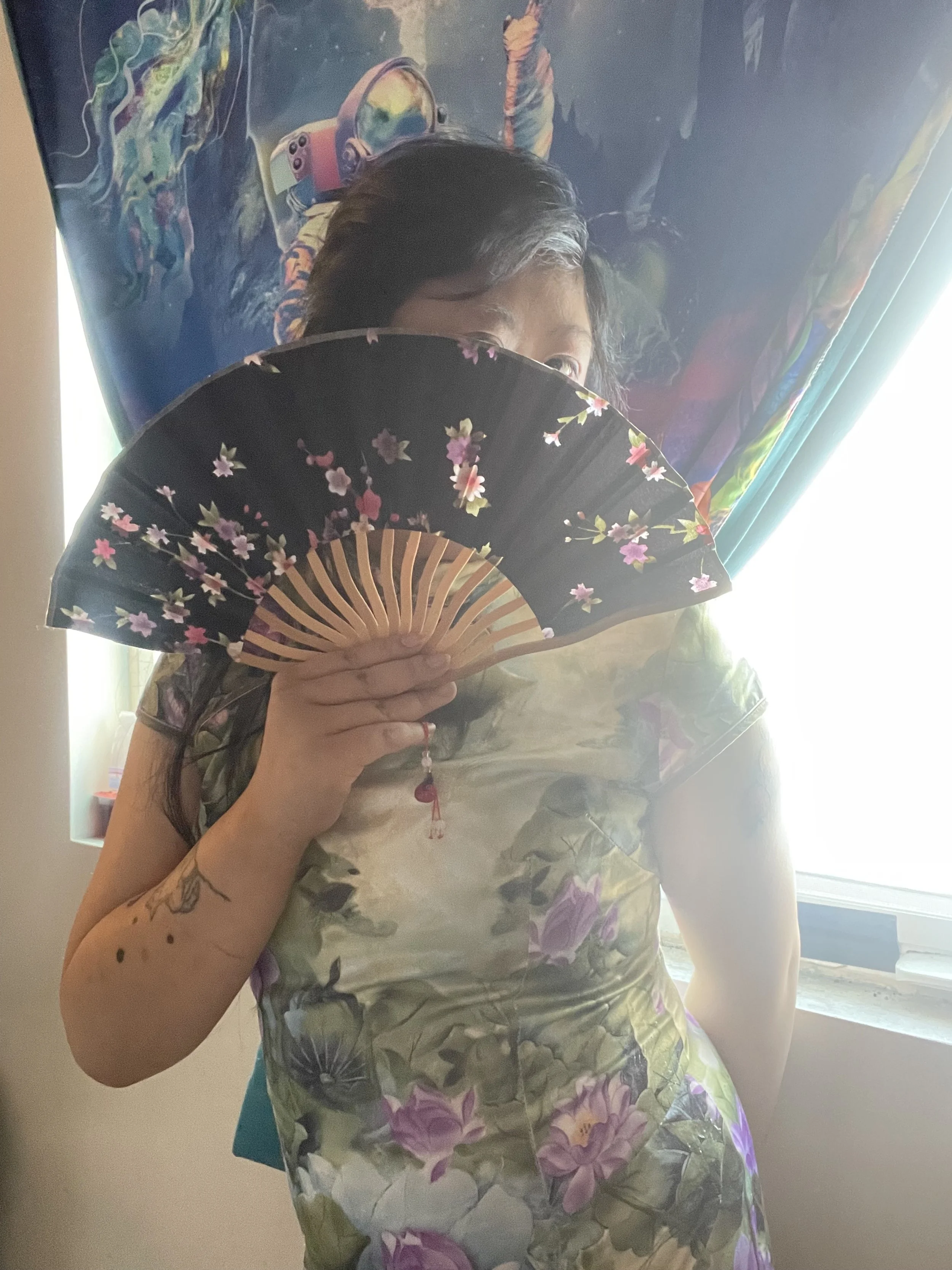 Woman wearing a floral dress, holding a black folding fan with pink and white flowers, partially covering her face, standing in front of a colorful curtain with an outer space design and a window with sunlight.