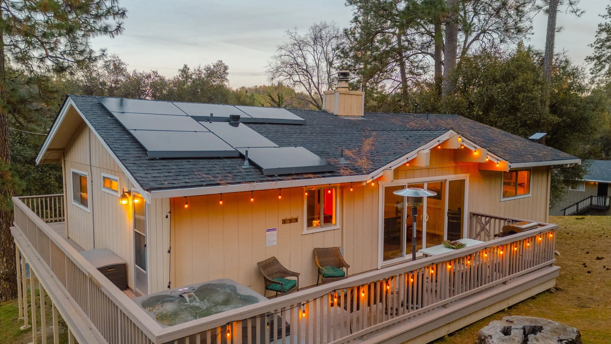 A house with a deck featuring string lights, two chairs with cushions, a hot tub, and solar panels on the roof, surrounded by trees at dusk.