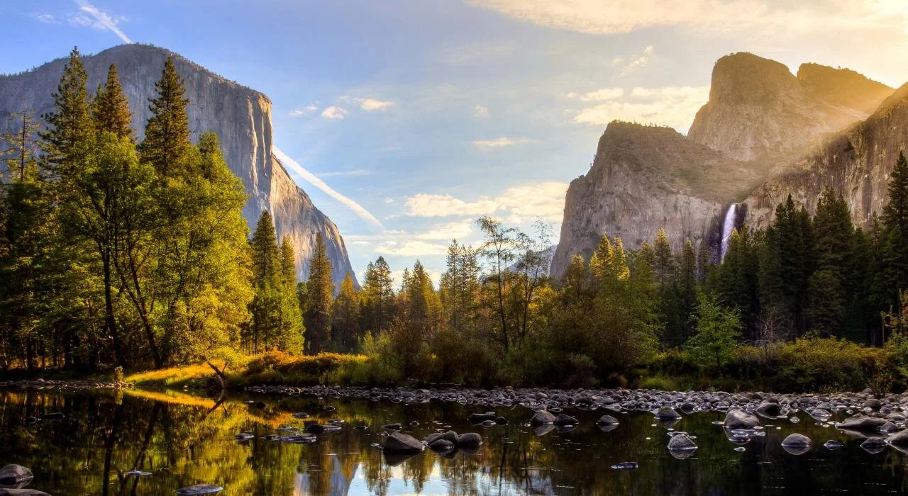A scenic view of a river flowing through a forest with tall trees, reflecting the surrounding mountains and sky during sunset, with waterfalls in the distance.