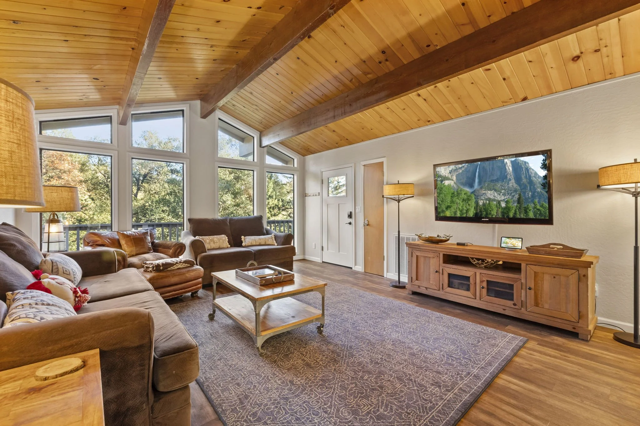 Living room with large windows, wooden ceiling, and wooden floor, furnished with sofas, a coffee table, a wooden TV stand, and a wall-mounted flat-screen TV showing a mountain scene.