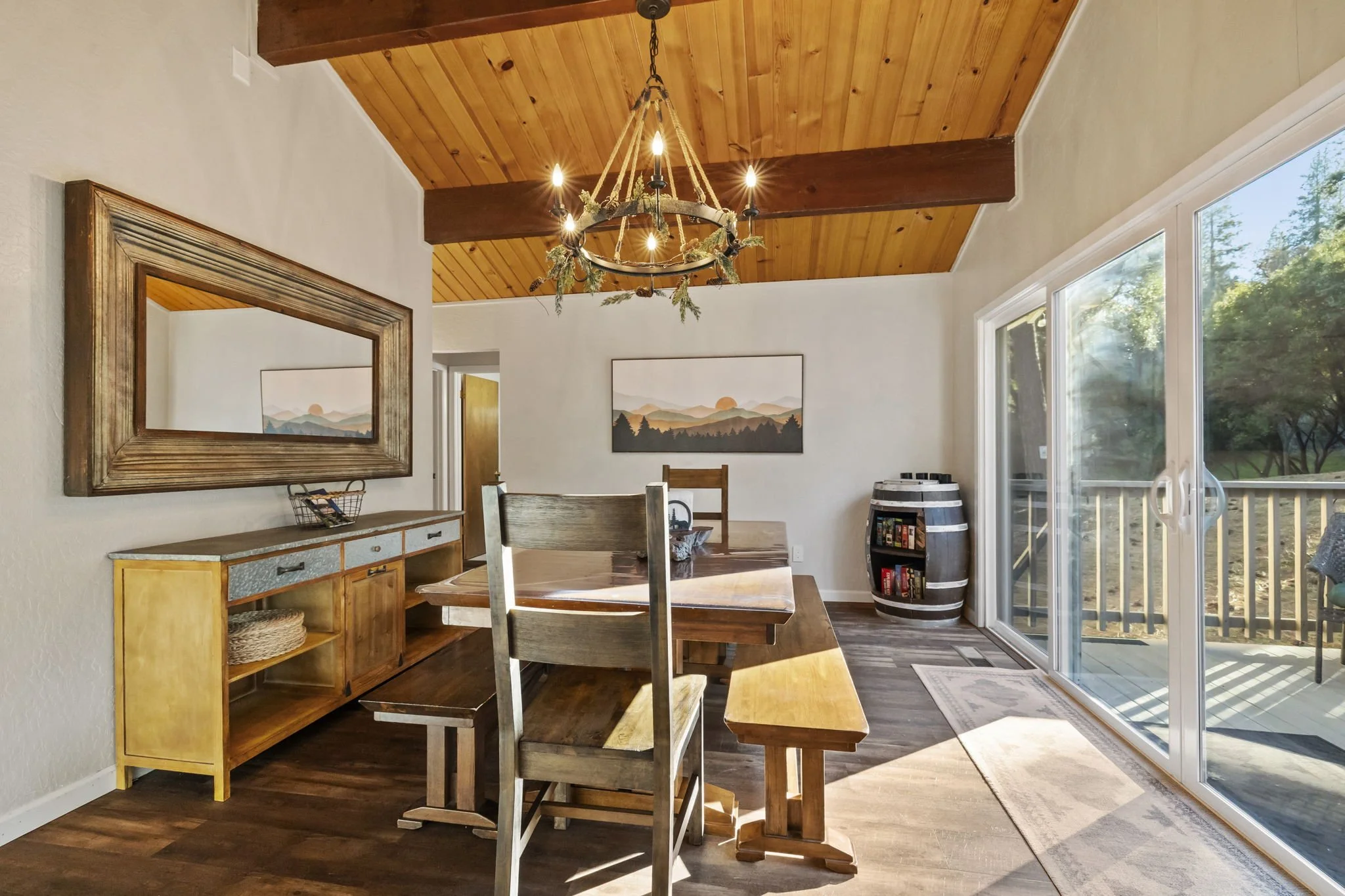 A dining room with a wooden table and chairs, a large mirror on the wall, a landscape painting, and a sliding glass door leading to a deck outside, with sunlight and greenery visible.