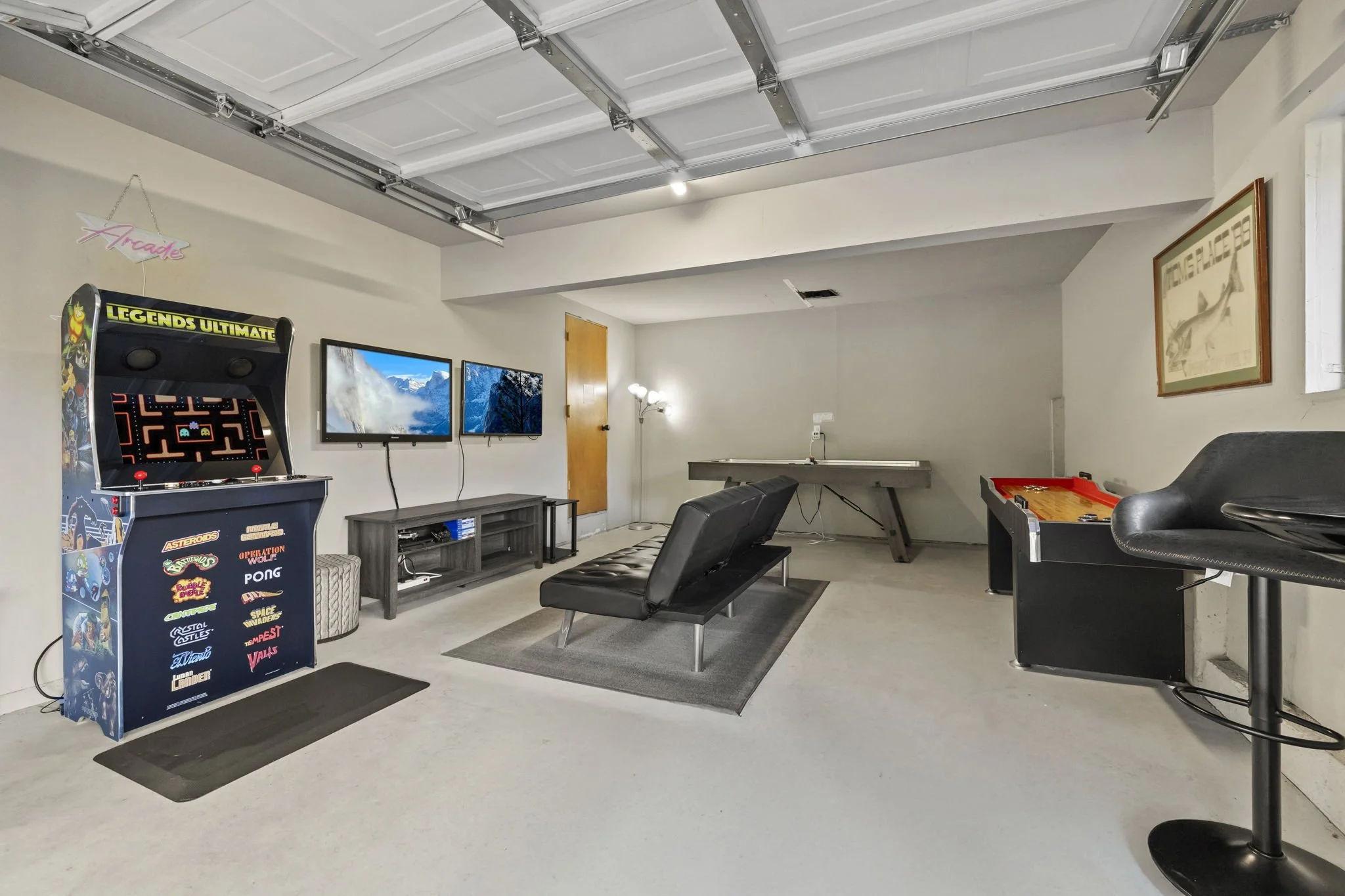A game room with a Legends Ultimate arcade game, two mounted TVs, a shuffleboard table, an air hockey table, a bar stool, a playstation 4, a floor lamp, and various posters and decorations.