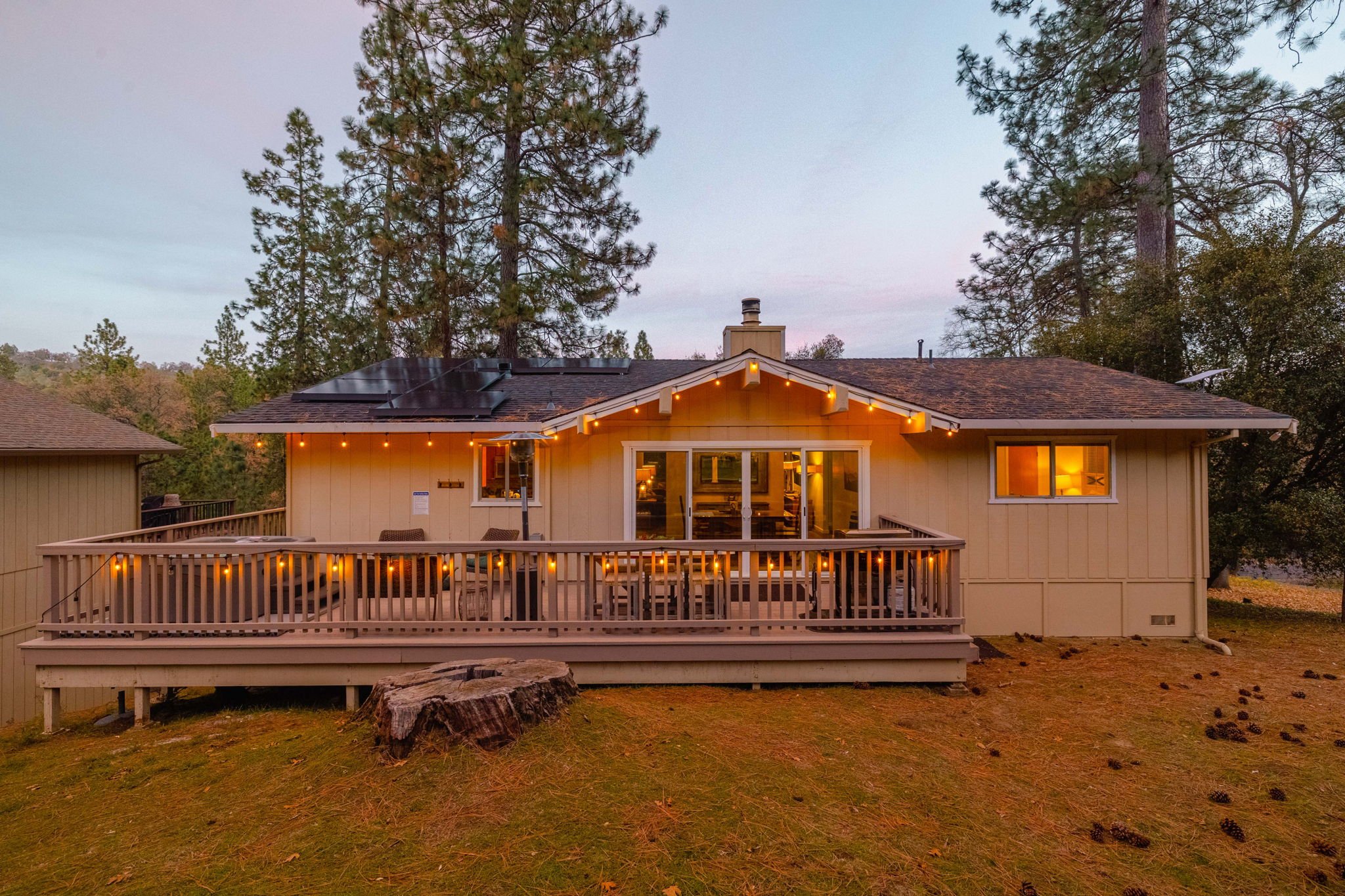 A house with a large wooden deck, string lights, and solar panels on the roof, surrounded by tall trees at dusk.