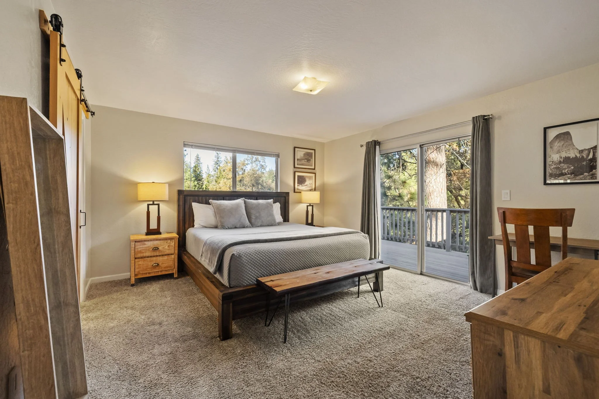 A bedroom with a large bed, wooden furniture, two bedside lamps, and sliding glass doors leading to a balcony with trees outside.
