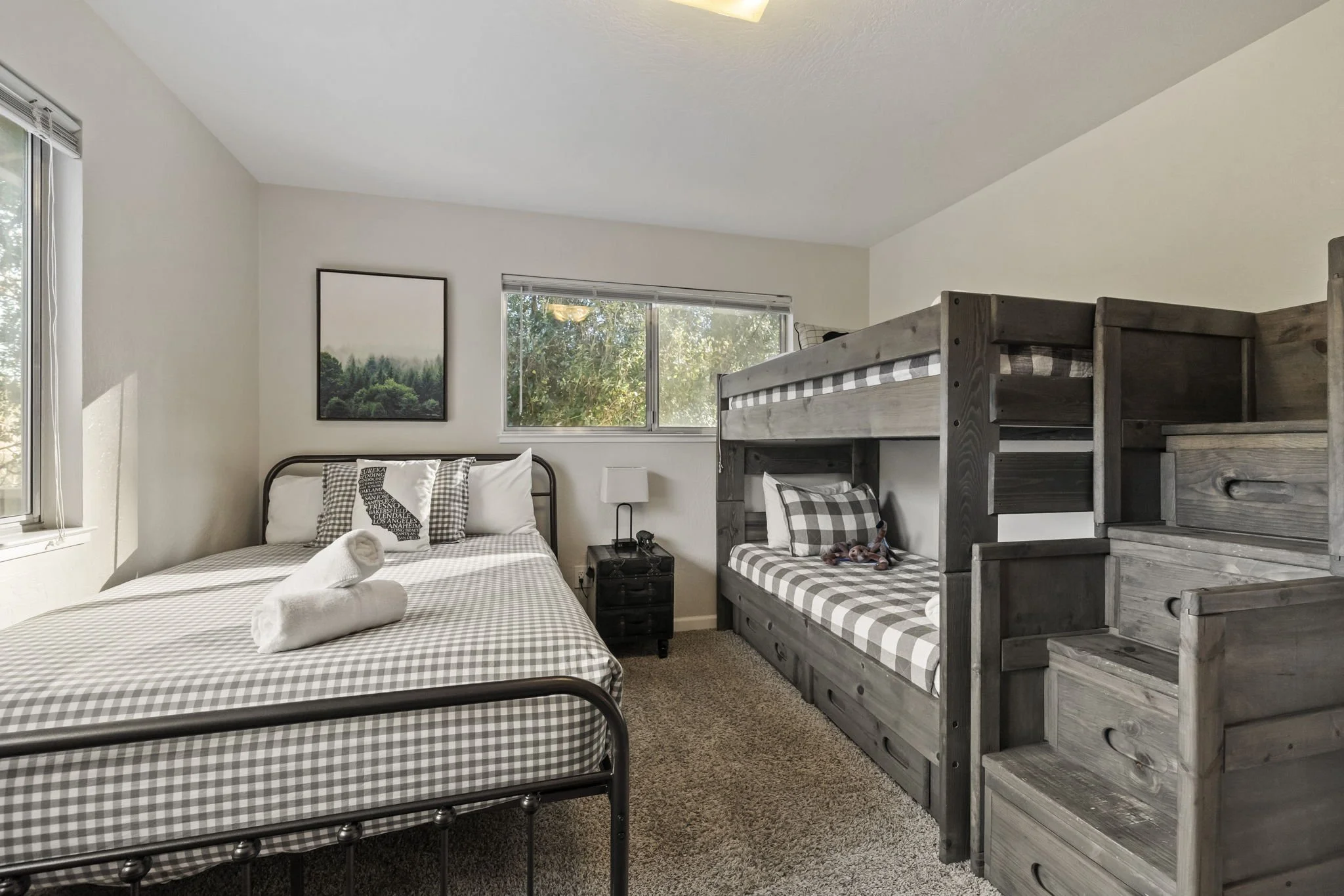 A bedroom with a full-size bed, a bunk bed with staircase, a nightstand, and two windows with natural light.