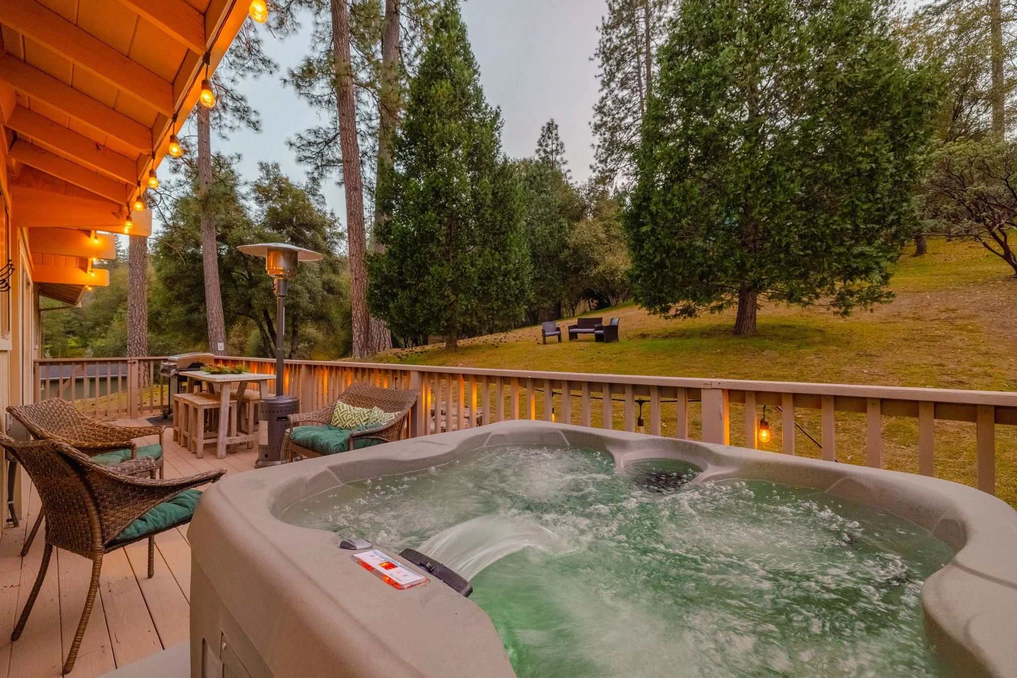Outdoor deck with hot tub, wicker chairs, a small table, string lights, patio heater, and scenic wooded landscape with trees and benches.