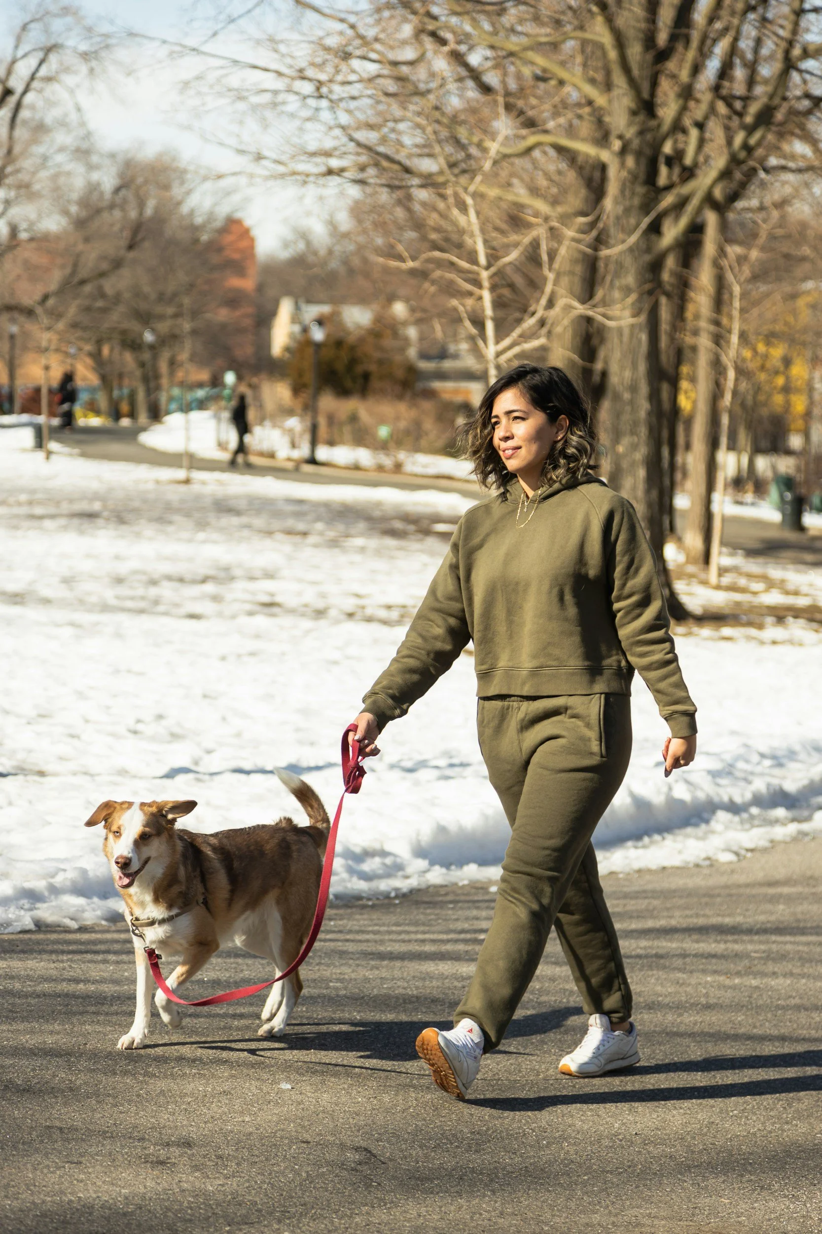 A woman in an olive green tracksuit walking her dog along a paved path in a park during winter, with snow on the ground and leafless trees in the background.