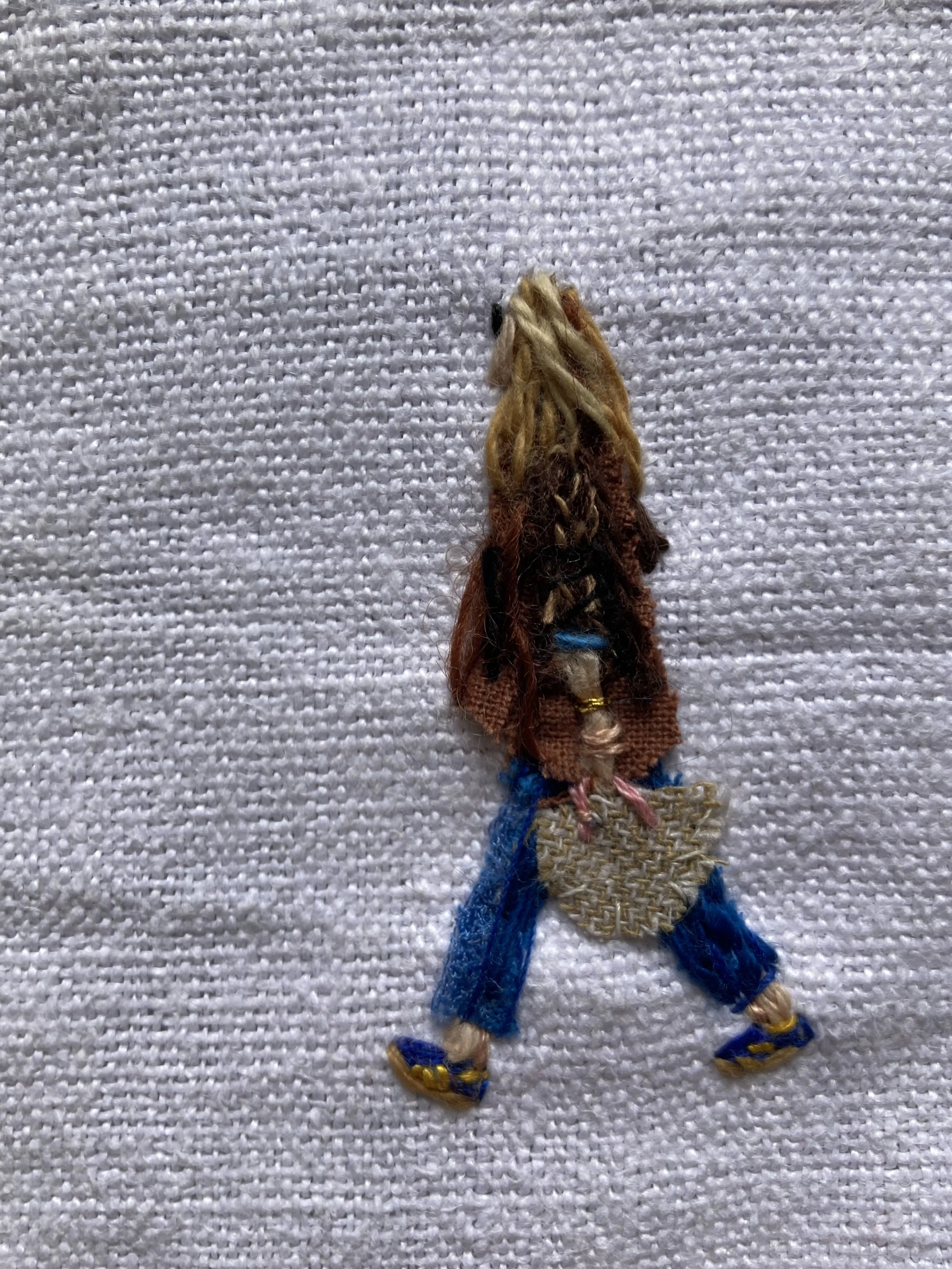 A textile depiction of a woman walking, shown from the back, with a beige woven bag and wearing blue pants, sandals, and a brown top.