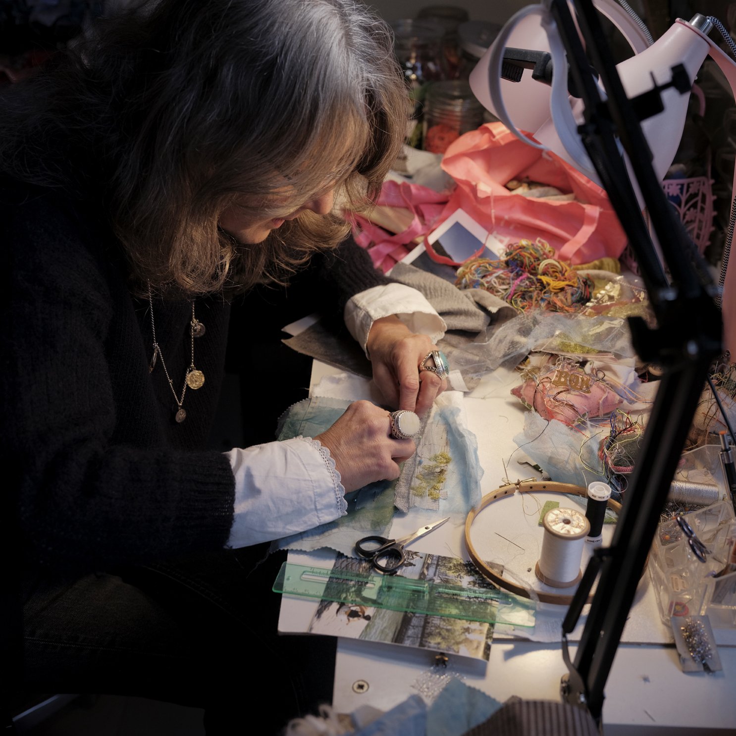 A woman working on an arts and crafts project at her cluttered workspace, surrounded by sewing materials, threads, scissors, and fabric.