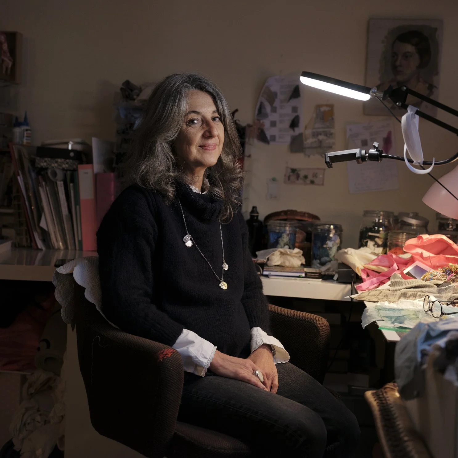 A woman with gray, wavy hair, wearing a black sweater, sitting in a cluttered workspace or studio, with arts and crafts materials on the table and shelves behind her.