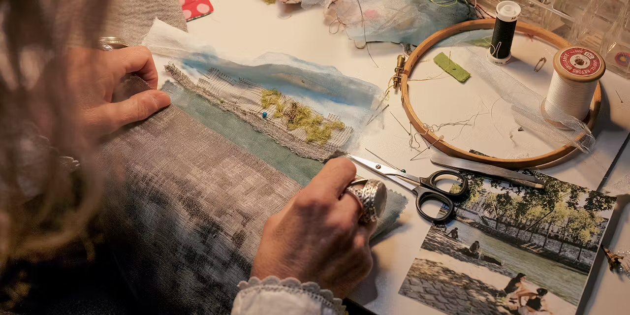 A woman is doing embroidery on a fabric at a sewing table with scissors, pins, thread, and a photo of people by a river nearby.
