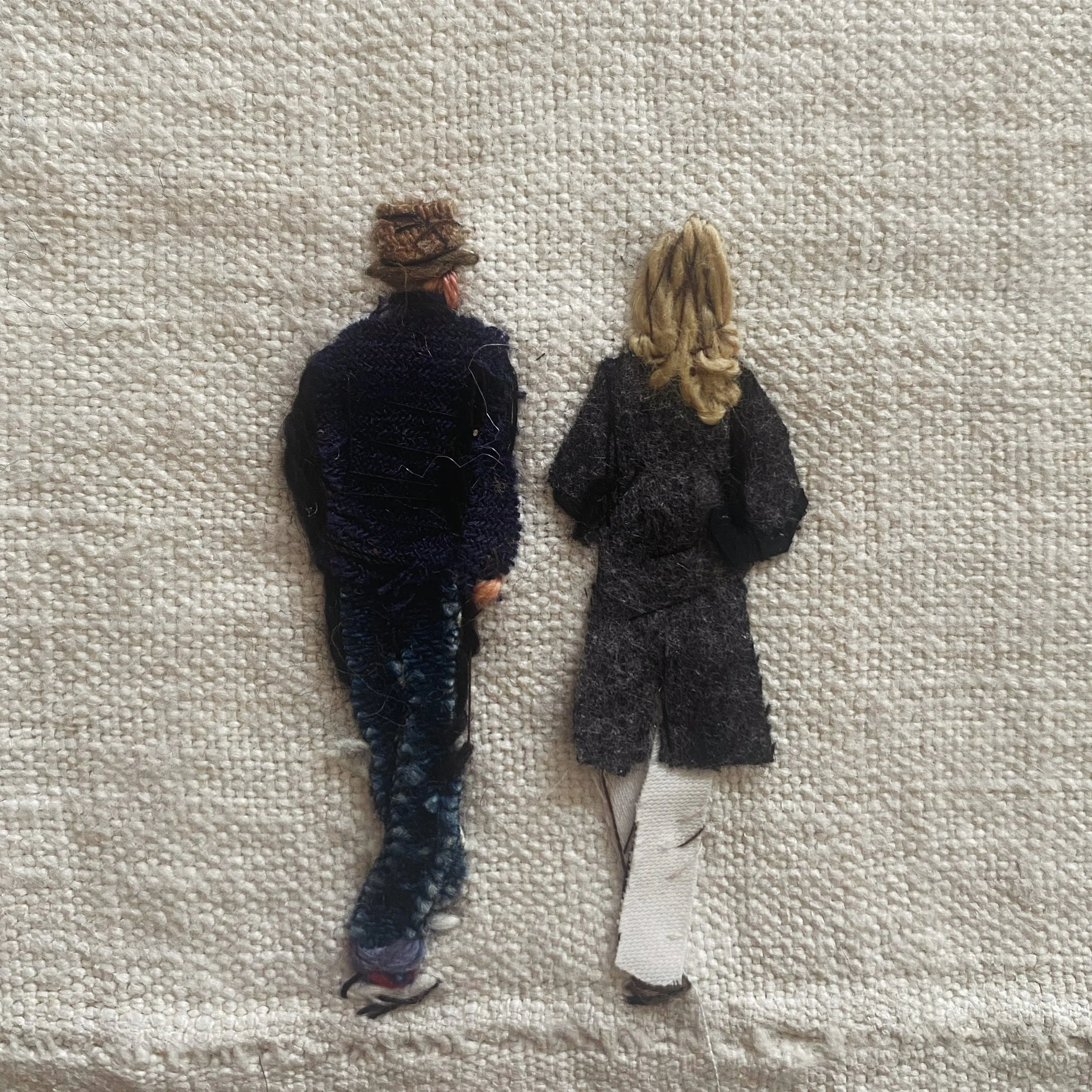 Two embroidered miniature figures of people, viewed from behind, lying on a textured fabric surface.