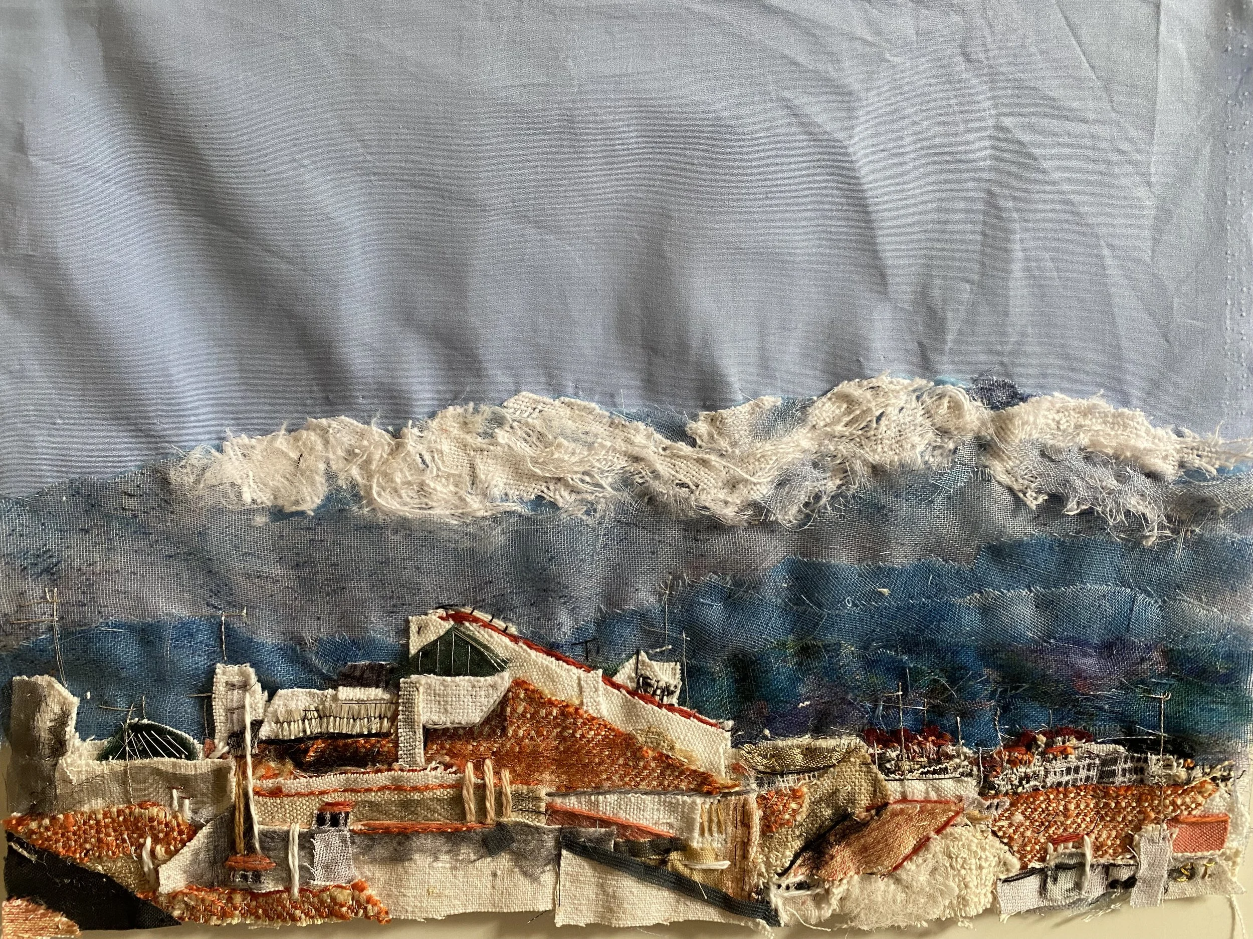 Textured fabric art depicting a landscape with clouds, mountains, and buildings in earthy and sky tones.
