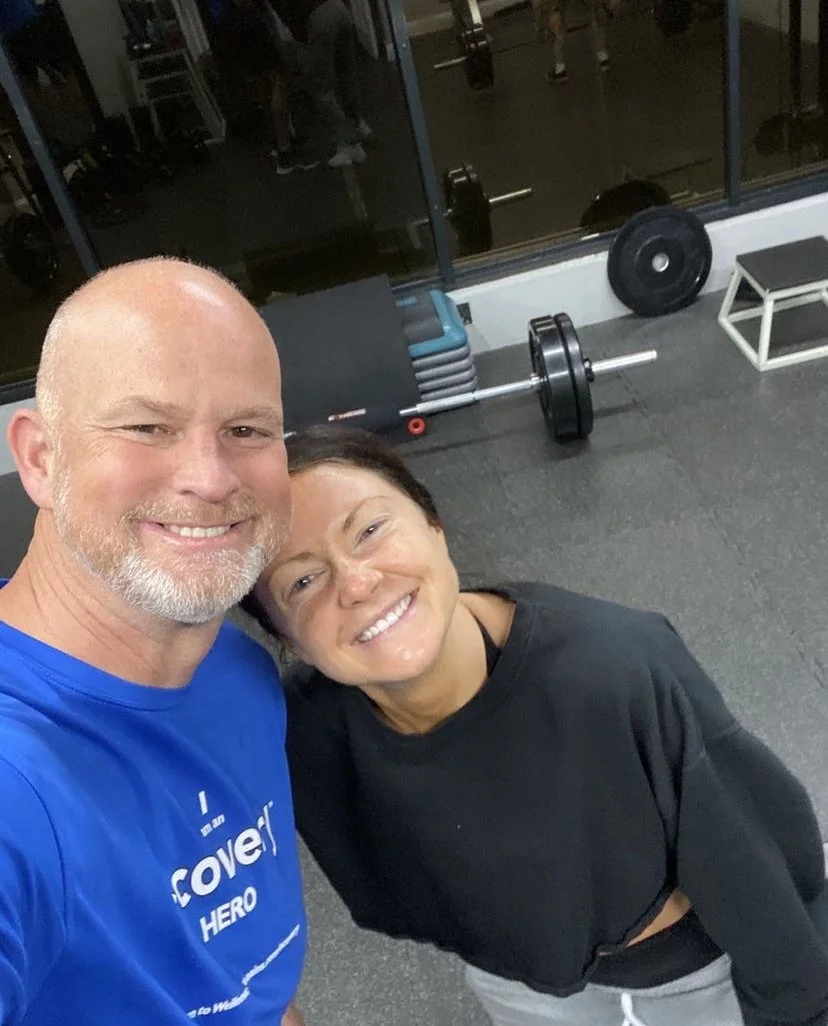 A man and woman smiling for a selfie in a gym. The man is wearing a blue shirt that says 'I am a mother HERO', the woman is in black, and both appear happy. Gym equipment, including a barbell, weights, and a step, are visible in the background.