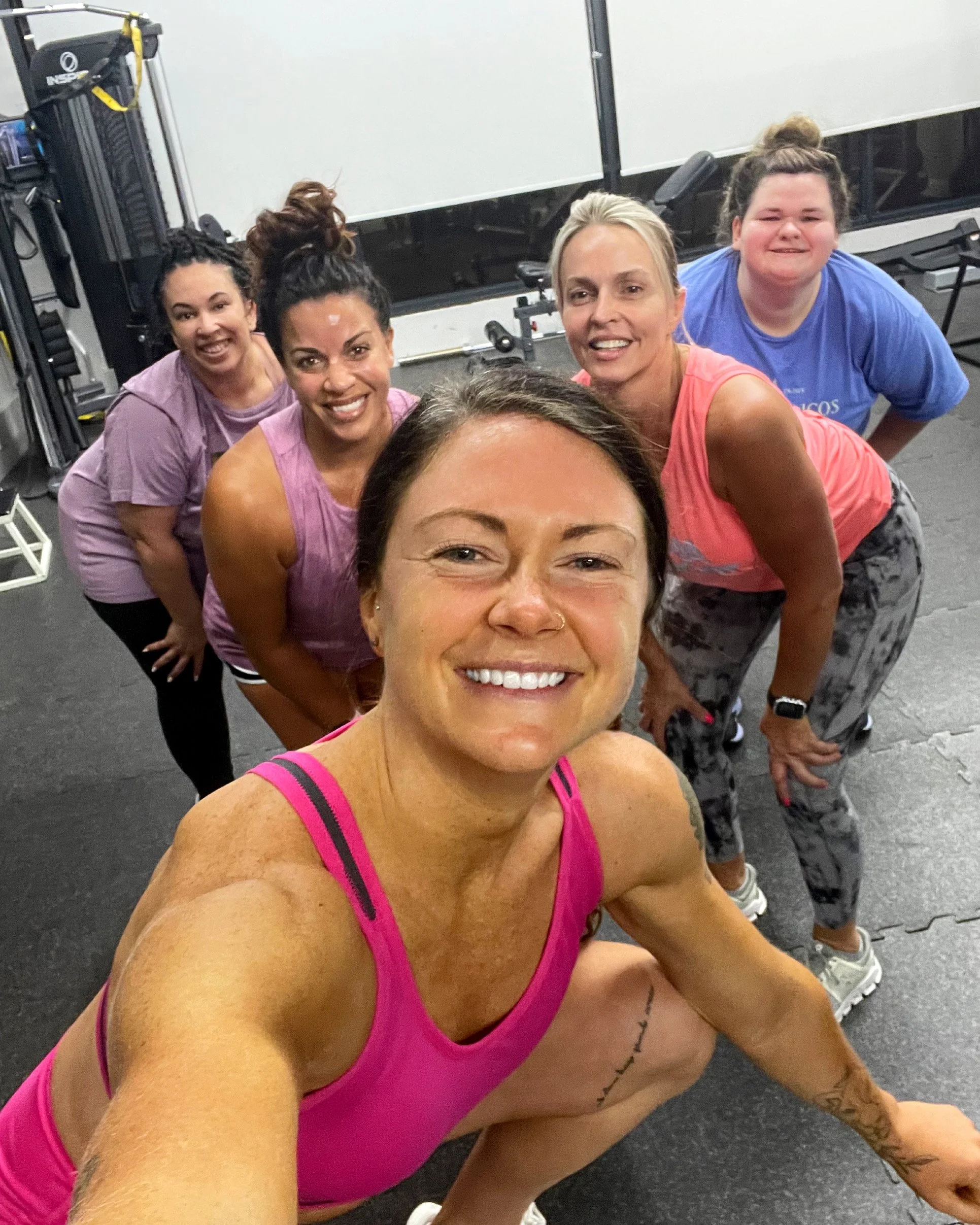 Group of women in workout clothes smiling inside a gym, taking a group selfie.
