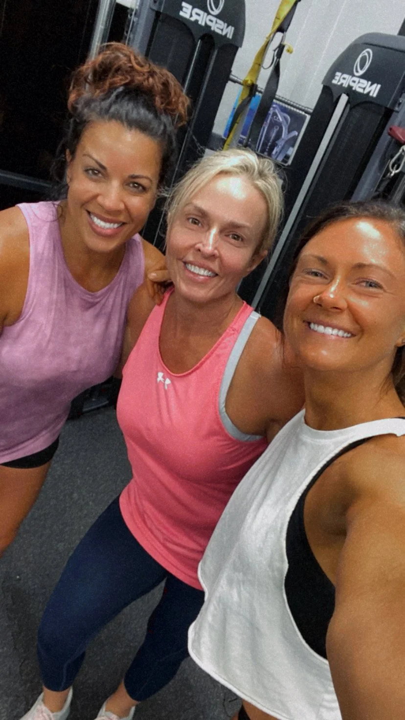 Three women taking a selfie at the gym, smiling, with fitness equipment in the background.