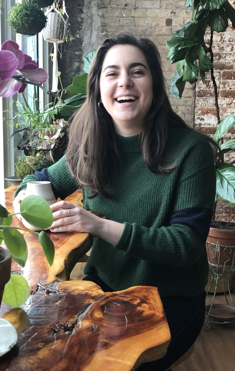 A smiling woman with long dark hair wearing a green sweater, sitting at a wooden table in a cozy, plant-filled indoor space with a brick wall background.