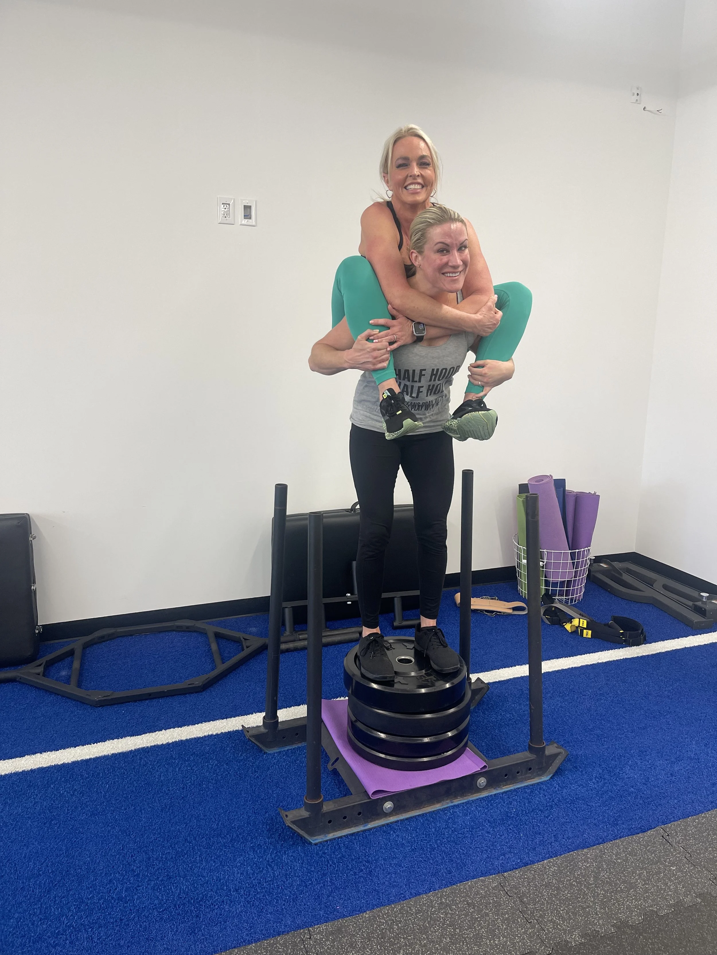 Two women exercising in a gym, one woman standing on a platform holding the other woman on her shoulders, both smiling. The woman on top is in a yoga pose, wearing green leggings and a black sports bra, and the woman on the bottom is wearing black leggings and a grey t-shirt. The gym has blue flooring, exercise equipment, and yoga mats in a wire basket.