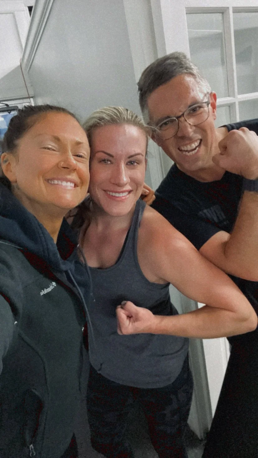 Three smiling people taking a selfie indoors, two women and one man, with the man making a fist and flexing muscles.