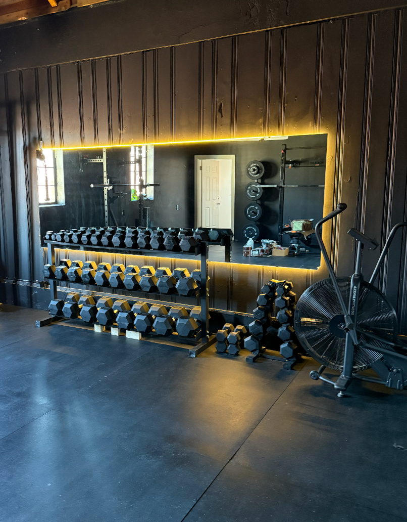 A gym with black walls, a large mirror, and a variety of workout equipment including dumbbells on racks, kettlebells on the floor, an exercise bike, and weight plates stacked vertically. The gym has a sleek, modern look with yellow accent lighting.