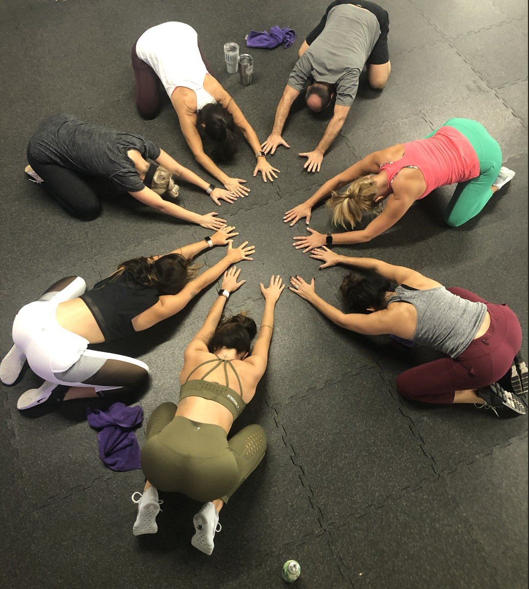 A group of eight people doing a yoga pose in a circle on the floor of a gym, with their hands and legs extended outward.