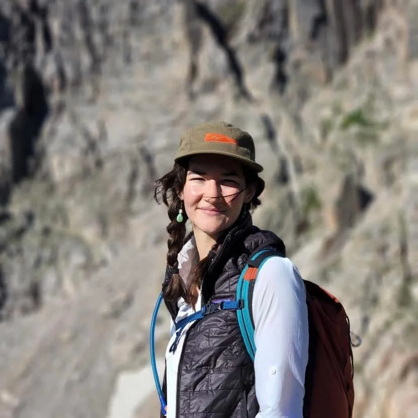 Young woman with braided hair smiling outdoors, wearing a brown hat, black vest over white shirt, and carrying a red backpack, with rocky mountain background.