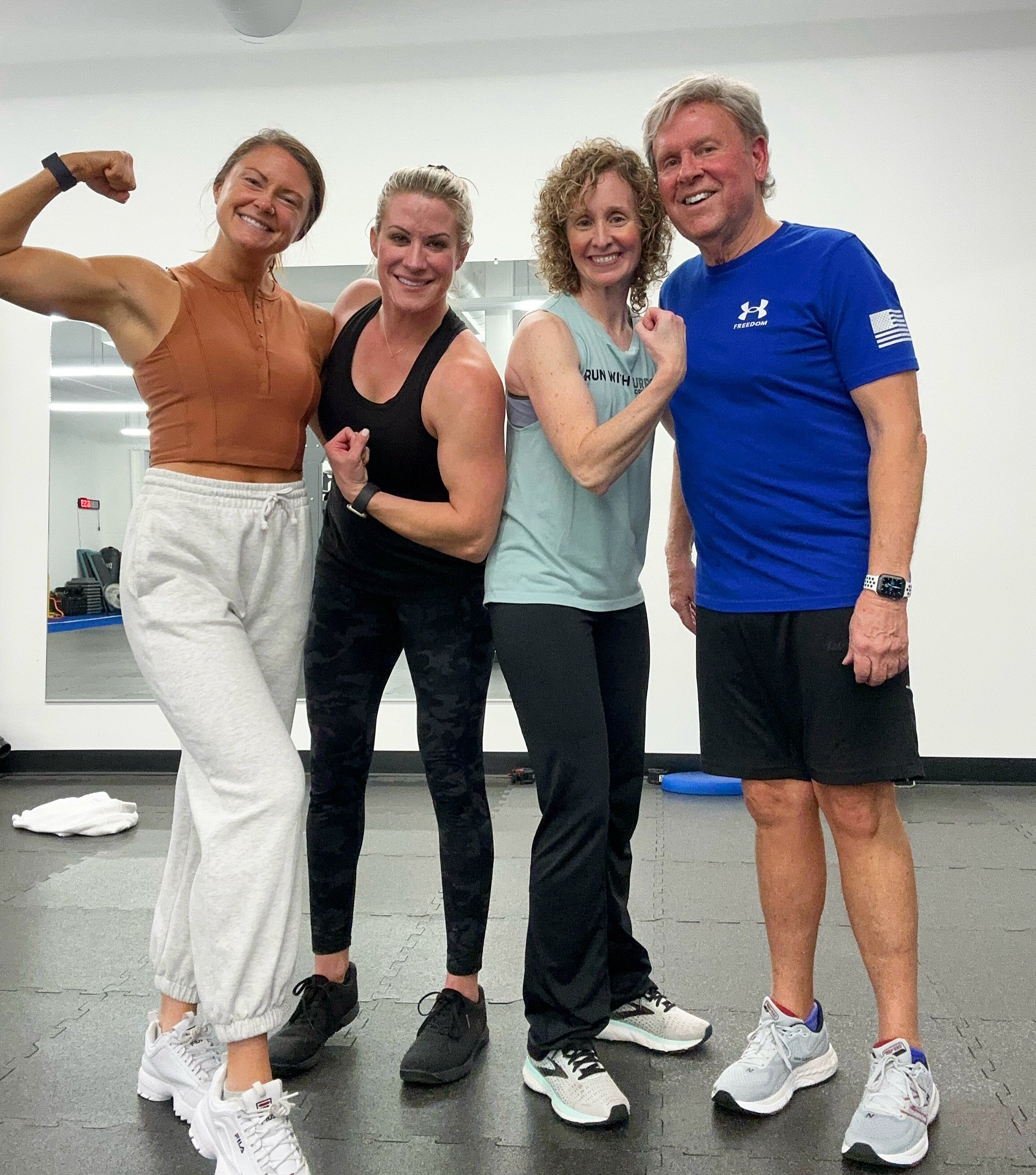 Four people in workout clothes standing together, smiling, and flexing their muscles in a gym.