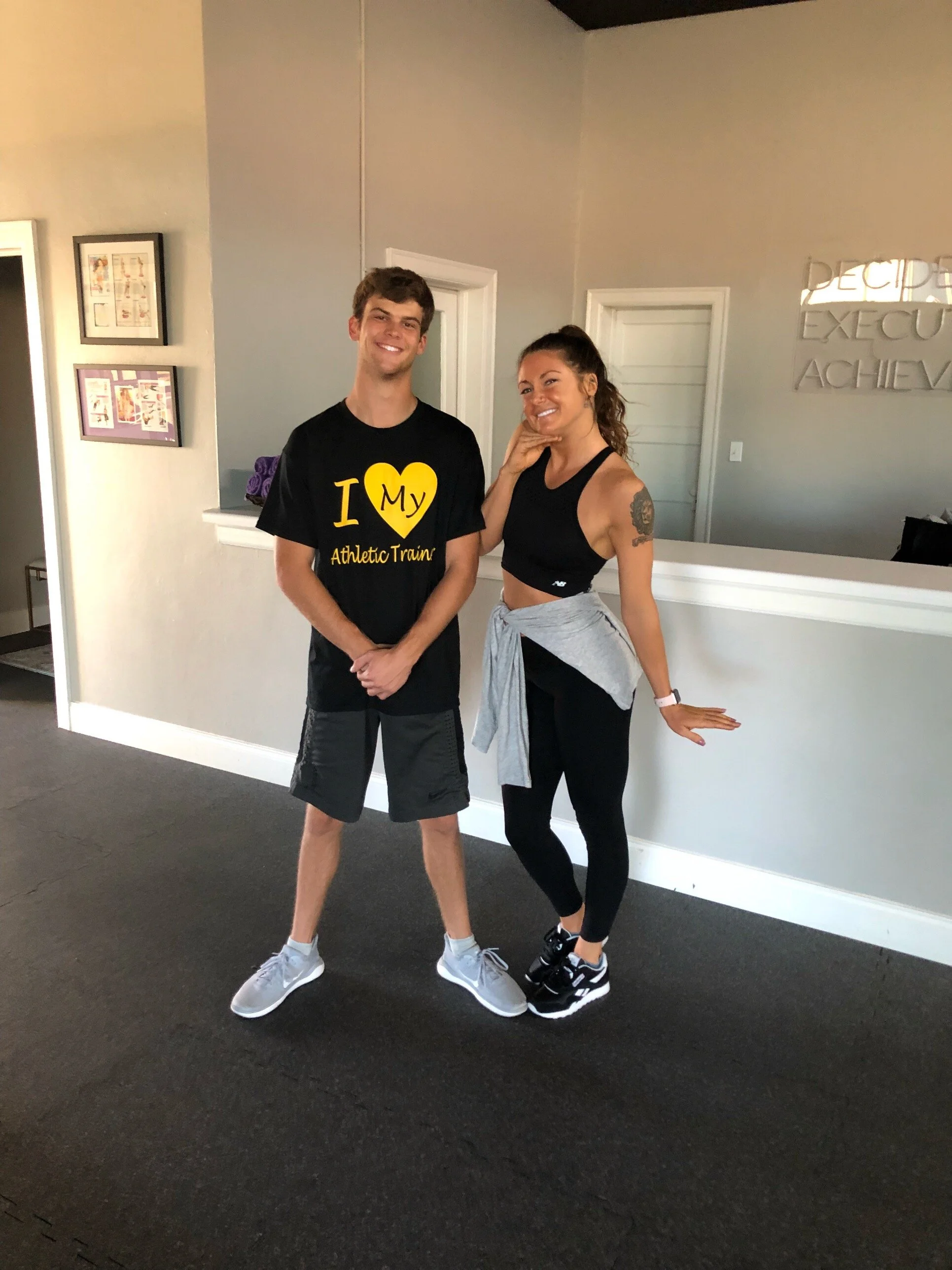 A man and woman are standing and smiling inside a gym. The man is wearing a black T-shirt with yellow text that says 'I Love My Athletic Trainer,' black shorts, and gray sneakers. The woman is wearing a black sports bra, black leggings, and black sneakers, with a light gray sweatshirt tied around her waist. She has a tattoo on her upper left arm.