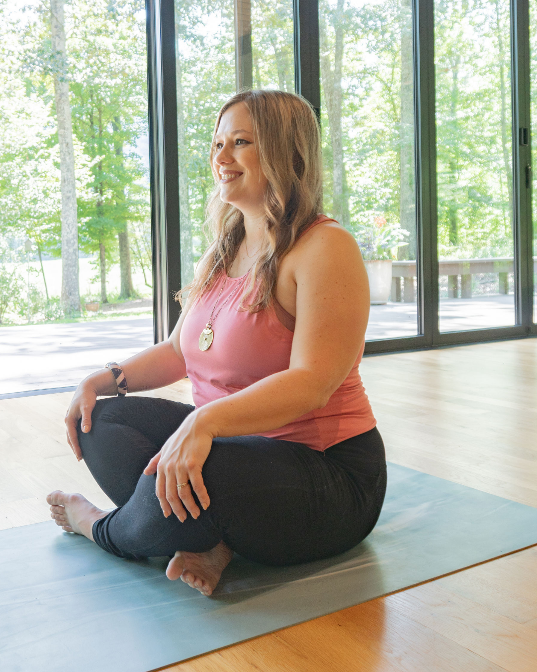 Woman meditating on a yoga mat inside, with large windows and a view of green trees outside.