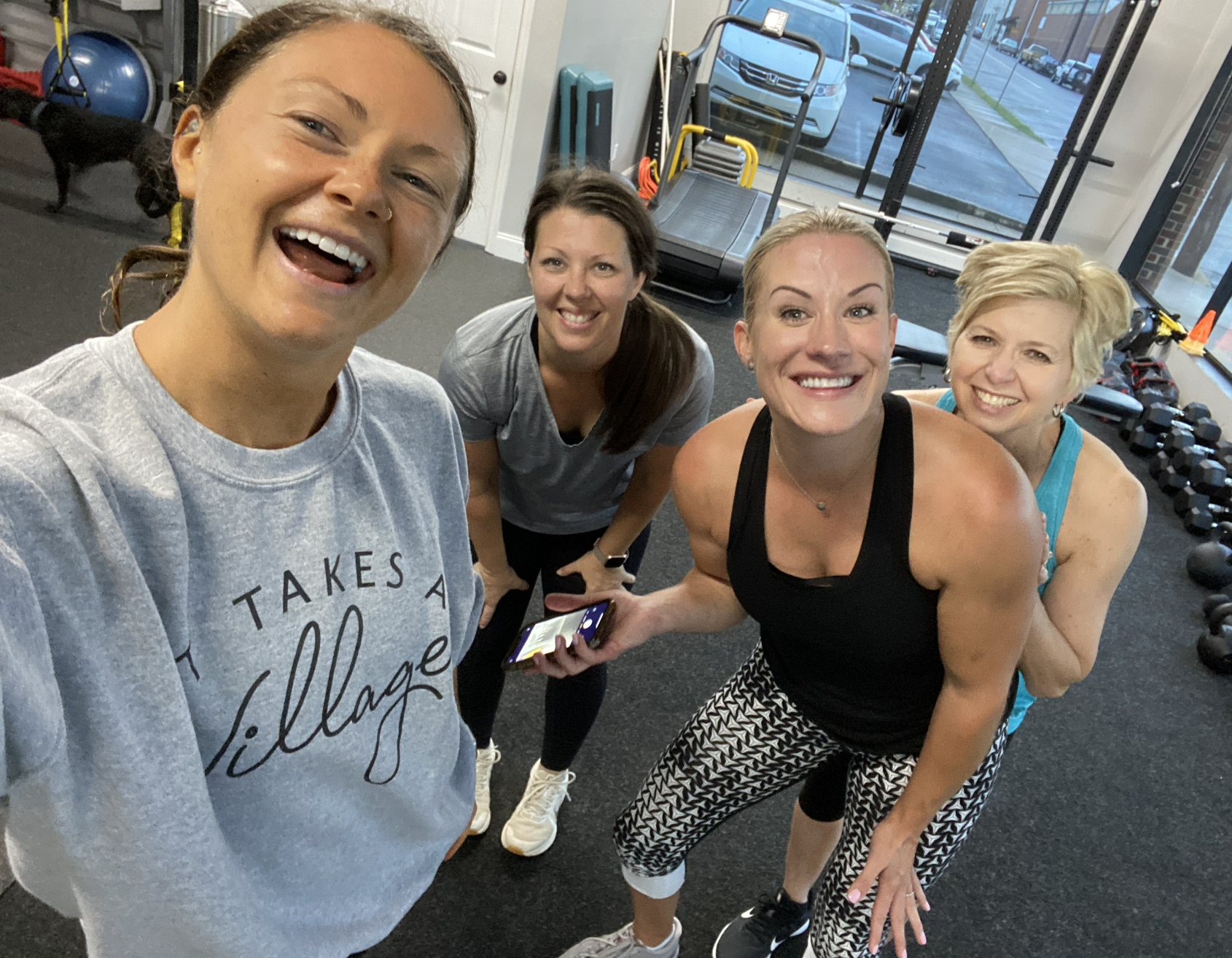 Four women in workout clothes smiling and taking a group selfie inside a gym, with workout equipment and a view of outside through large windows.