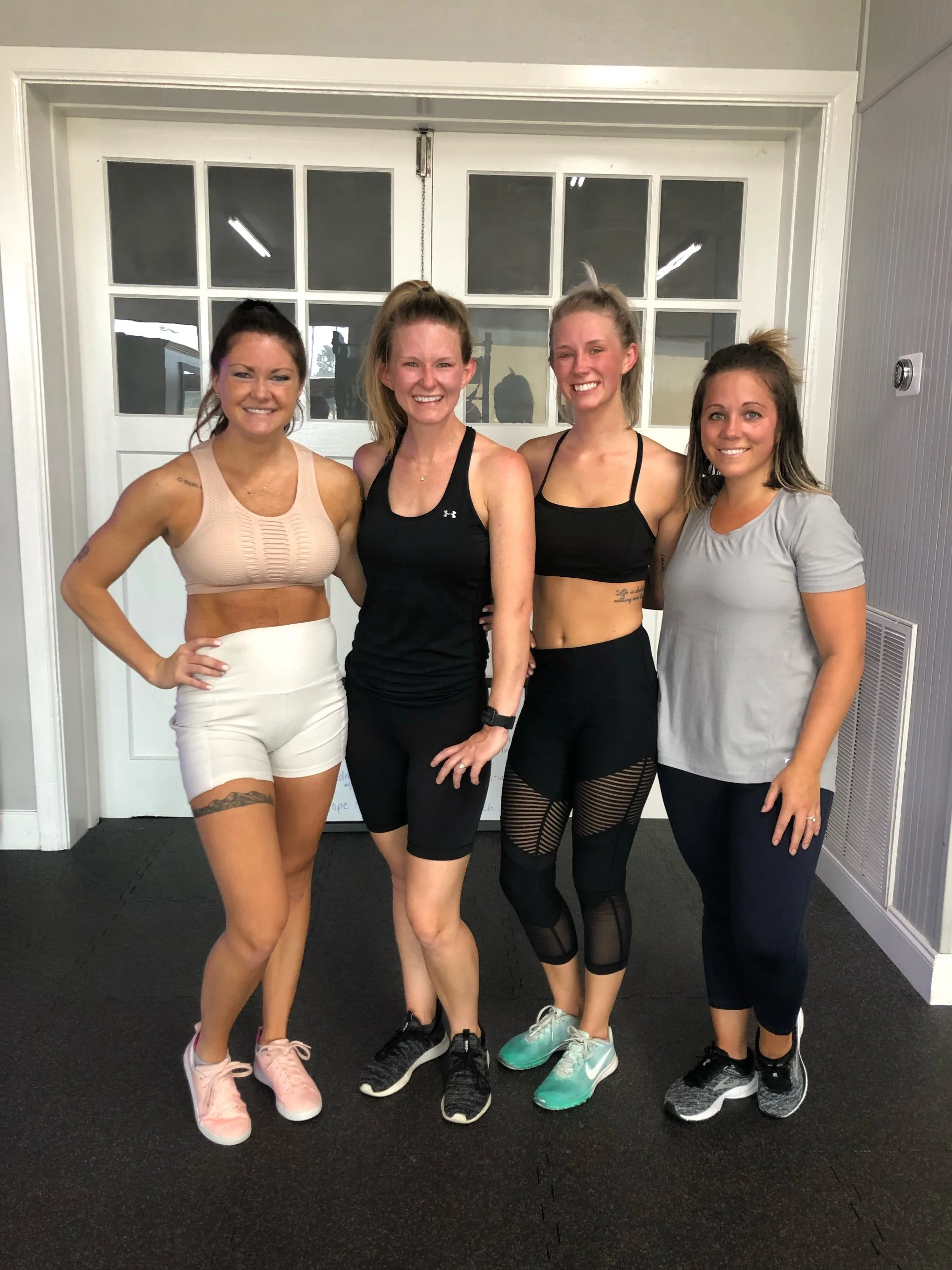 Four women in workout attire standing together inside a gym, smiling at the camera.