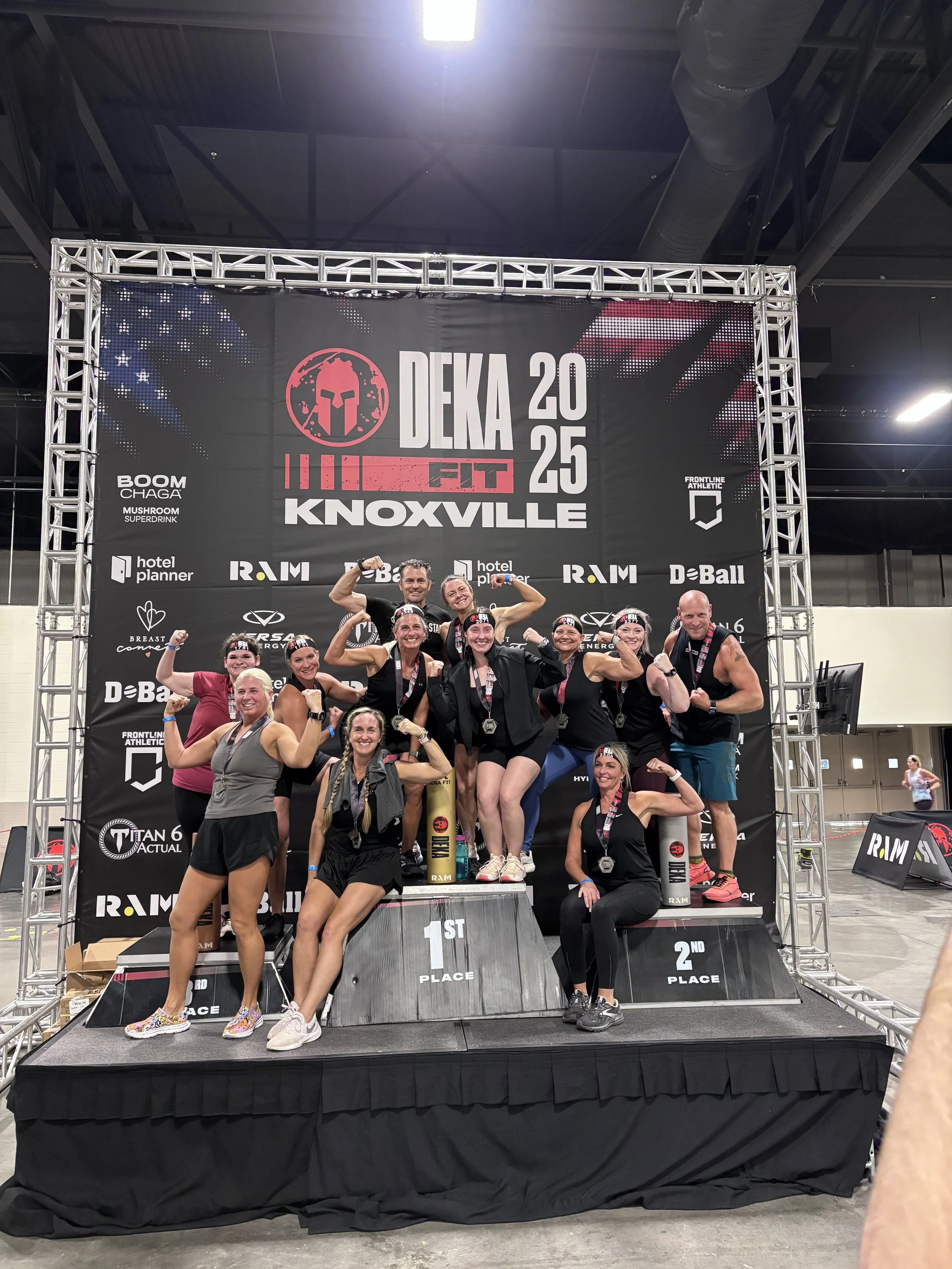 Group of women and one man on the winners' podium at a fitness competition, flexing their muscles and smiling, with medals around their necks, at the DEKA Fit Knoxville 2025 event.