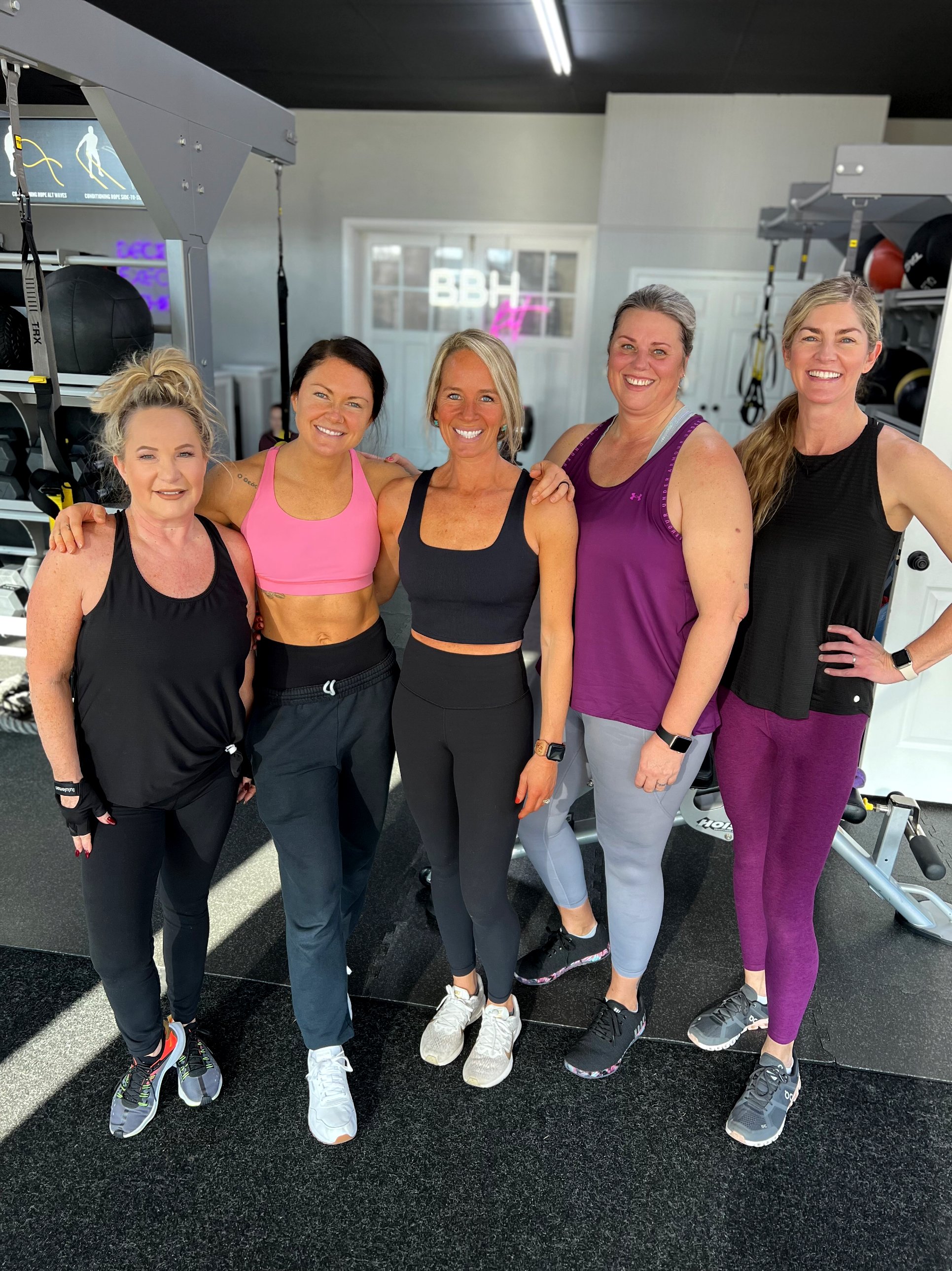 Five women in workout attire standing together in a gym, smiling at the camera.