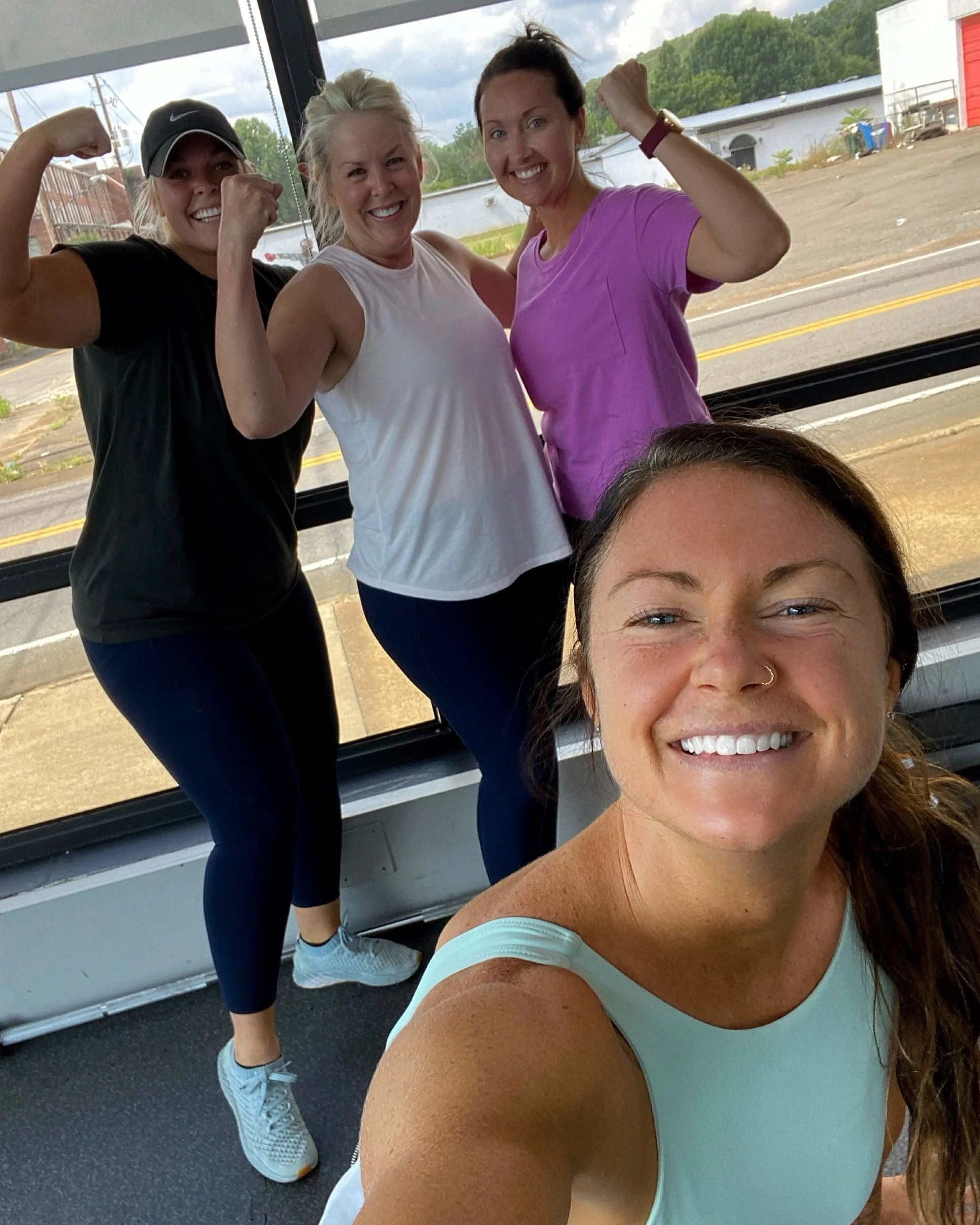 Four women smiling and flexing their arms inside a bus or train with large windows and a view of the outdoors.