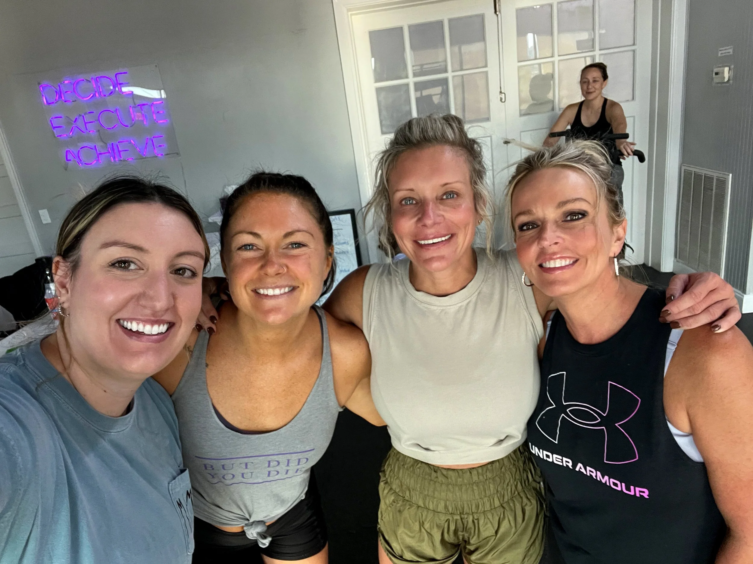 Group of five smiling women taking selfie, one woman in background on a treadmill, neon sign with motivational words in the background.