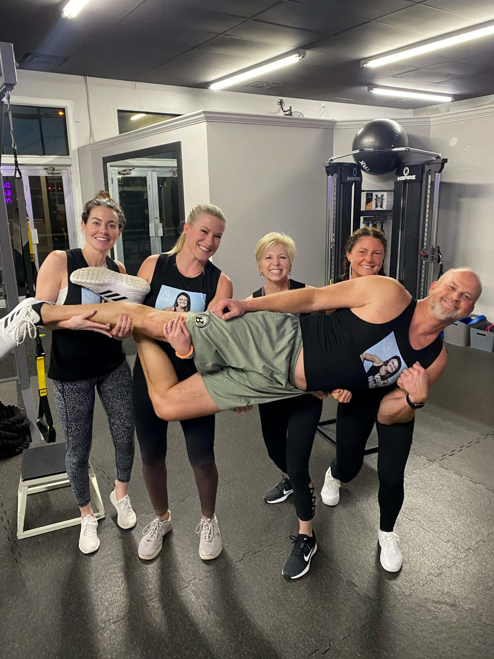 Four women lifting a man in a gym. The man is lying horizontally with his legs supported by one woman and his upper body held by others. Everyone is smiling and wearing workout attire.