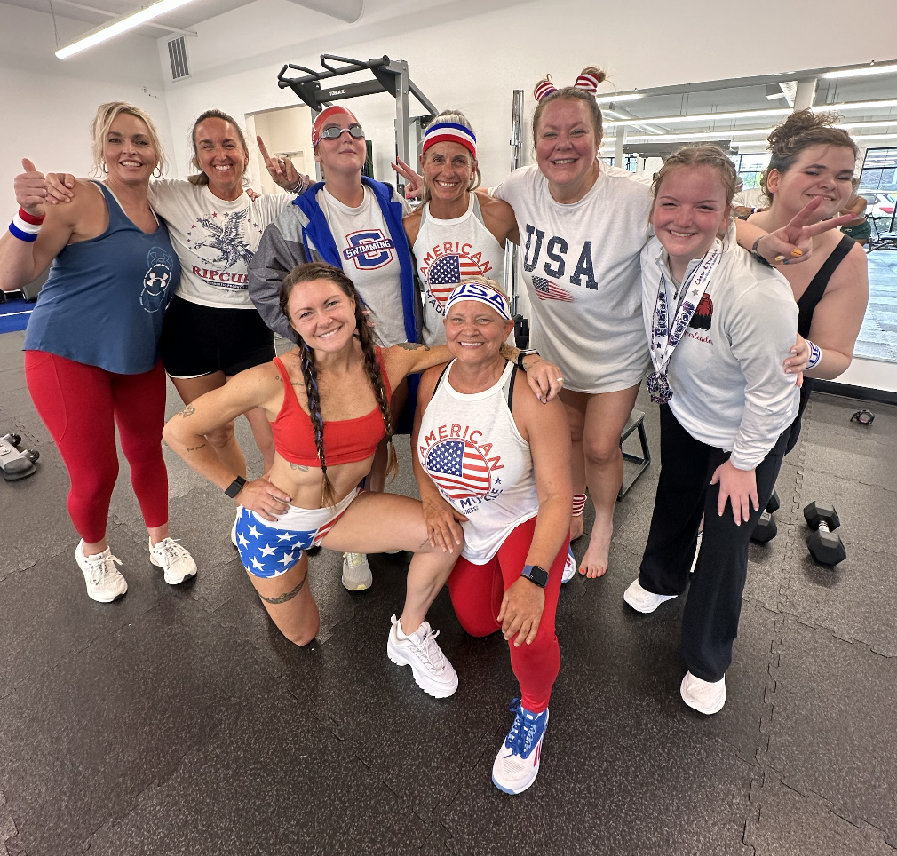 Group of nine women posing together in workout attire with American patriotic themes at a gym.