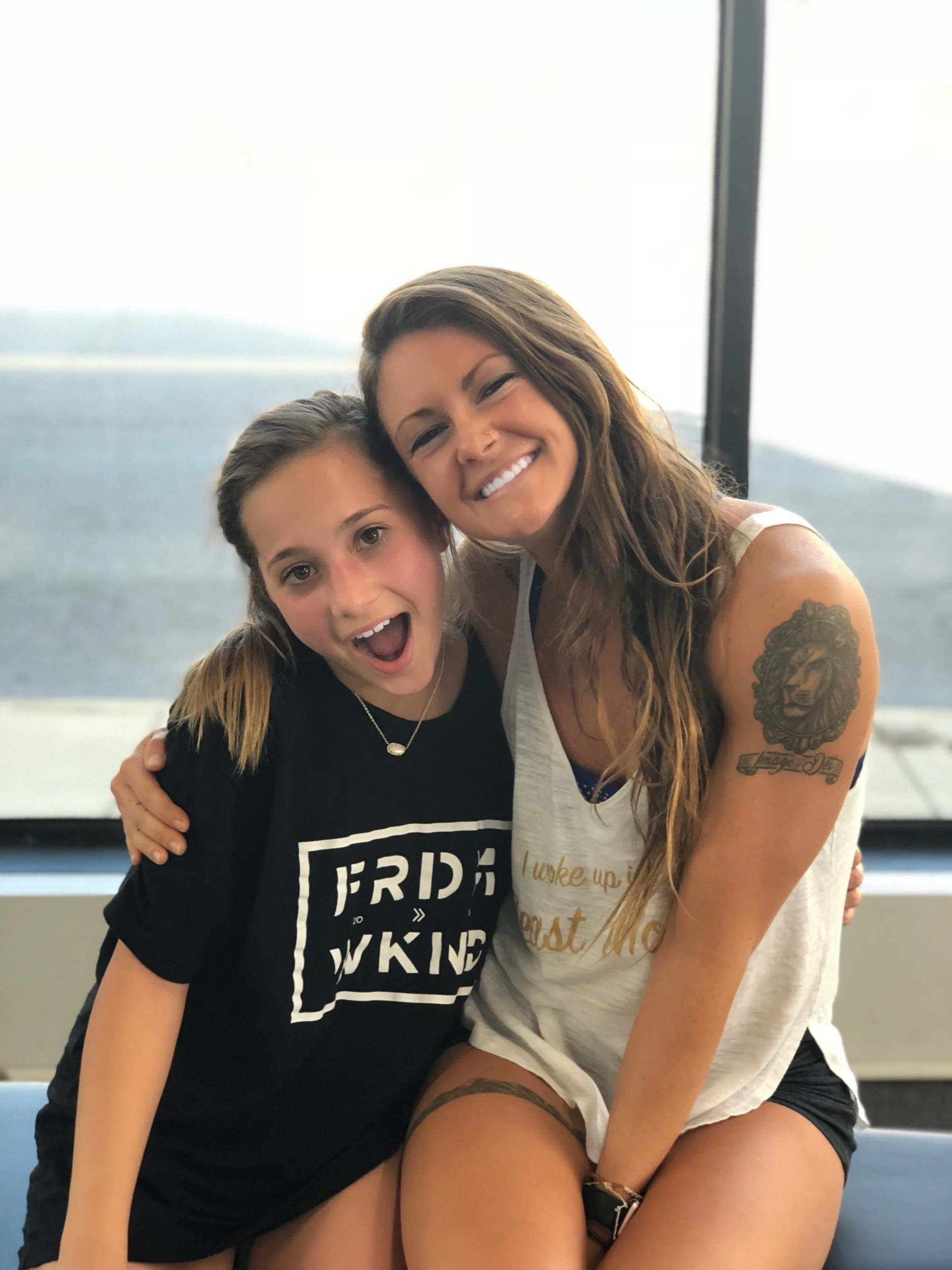 Two women smiling and hugging inside with a window showing mountains and sky in the background.