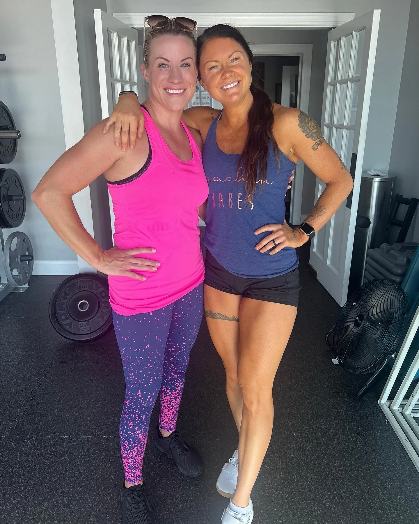 Two women smiling and posing for a photo inside a gym; one in a pink sports tank top and patterned leggings, the other in a blue tank top and black shorts.