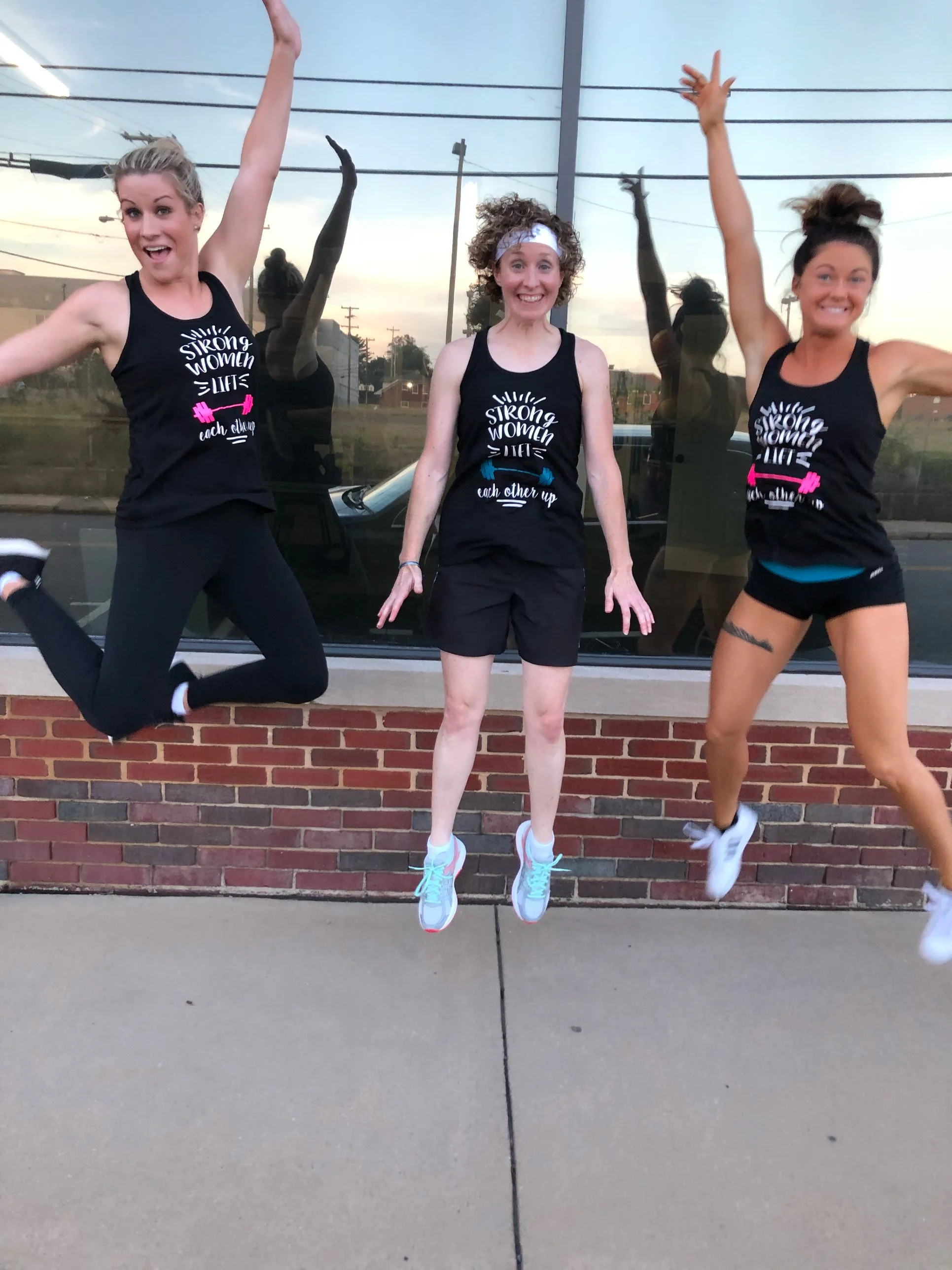 Three women in athletic clothing jumping in front of a window with a reflection of their jumping poses. They are wearing matching tank tops with the words 'Strong Women' and weights printed on them.