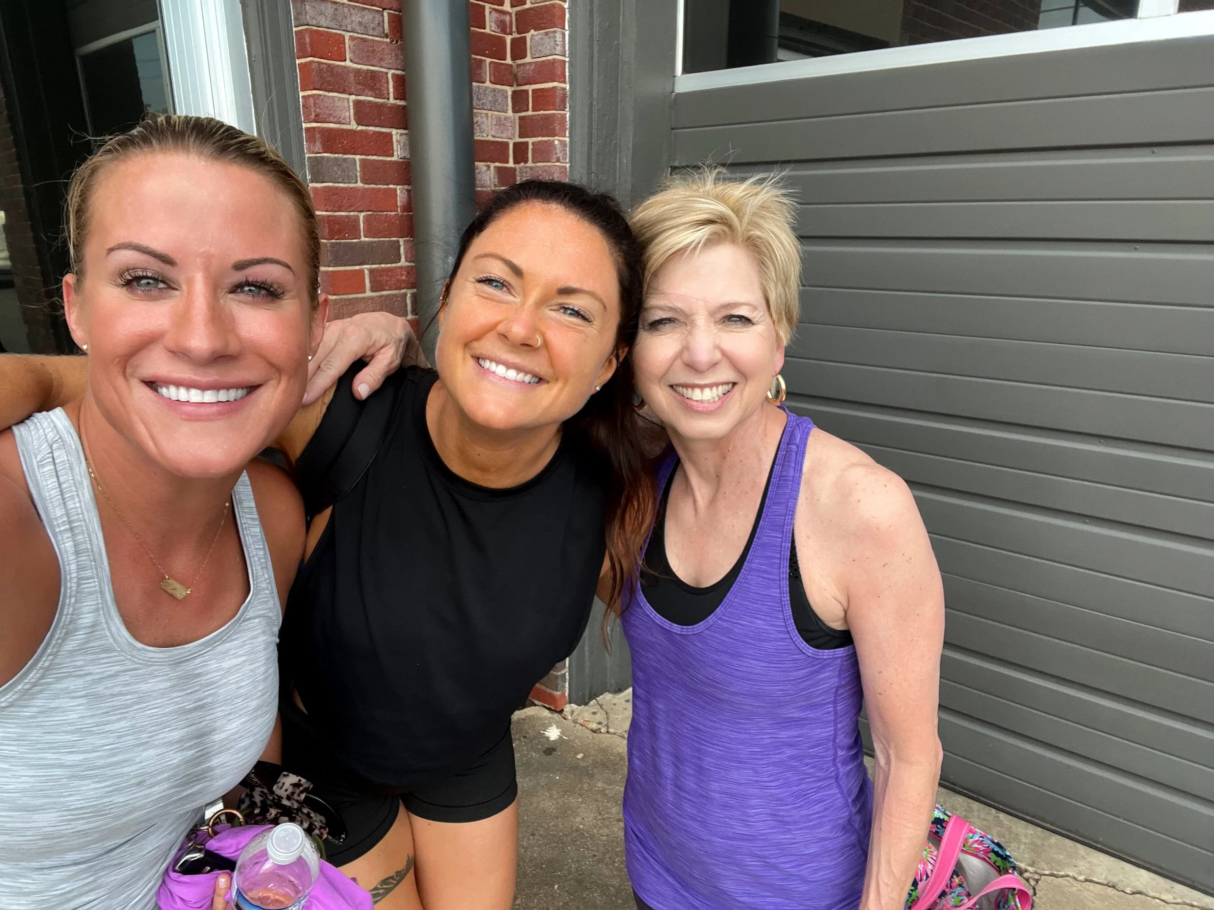 Three smiling women posing outdoors, one in a white top, one in a black top, and one in a purple top, with backpacks and water bottles, standing in front of a brick wall and grey shutter.