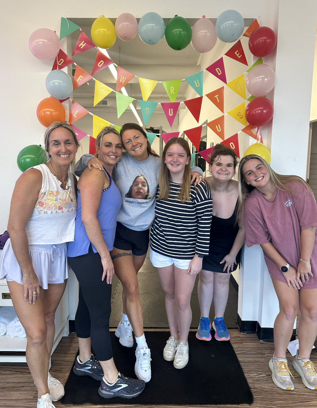 Group of six women and girls standing inside, celebrating with colorful balloons and bunting that says "Happy Birthday" and "How Are You" in the background.