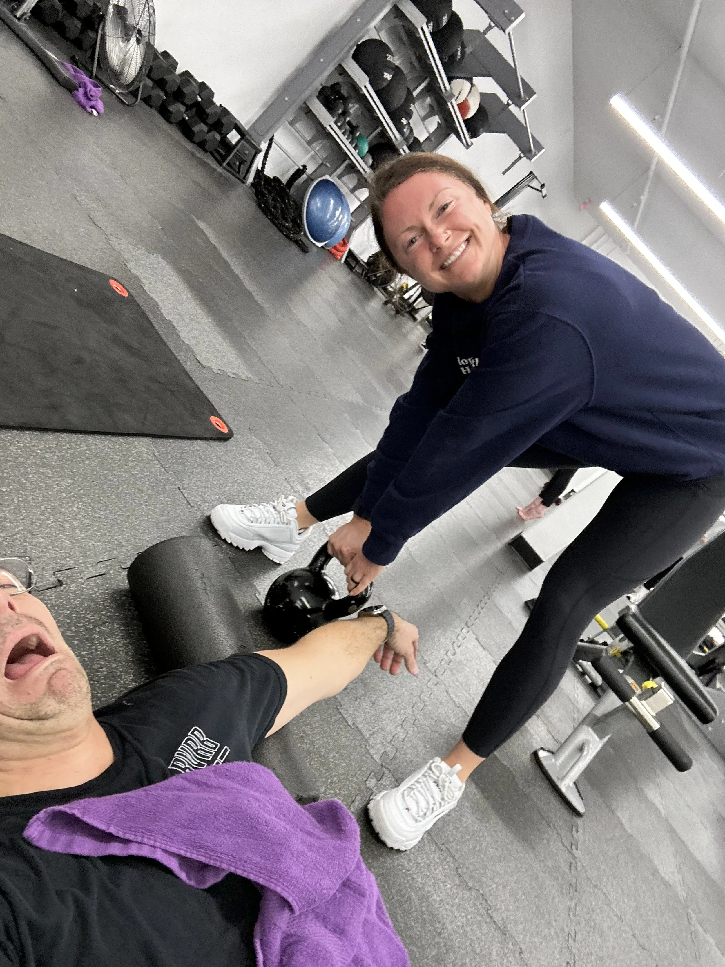 Two people in a gym, one lying on the floor holding a kettlebell, and the other assisting with the kettlebell, both smiling.