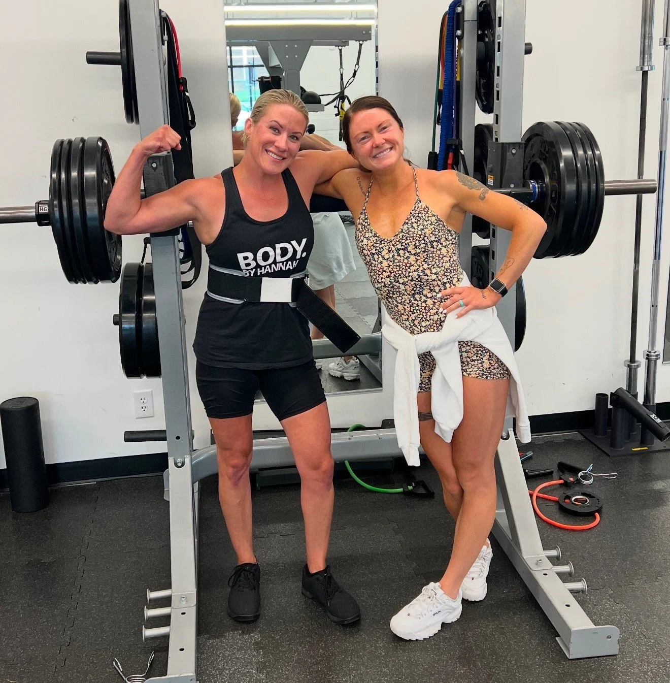 Two women at the gym, one in workout attire flexing her bicep, the other smiling with a white sweater tied around her waist, standing next to a barbell on a squat rack.