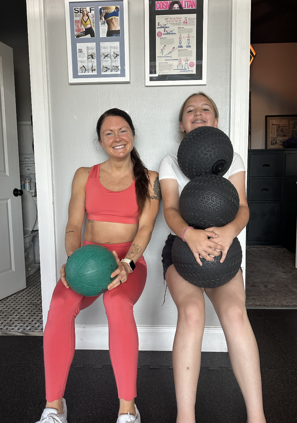 Two women at a gym, one sitting and holding a green exercise ball, the other standing and holding three black textured exercise balls, both smiling.