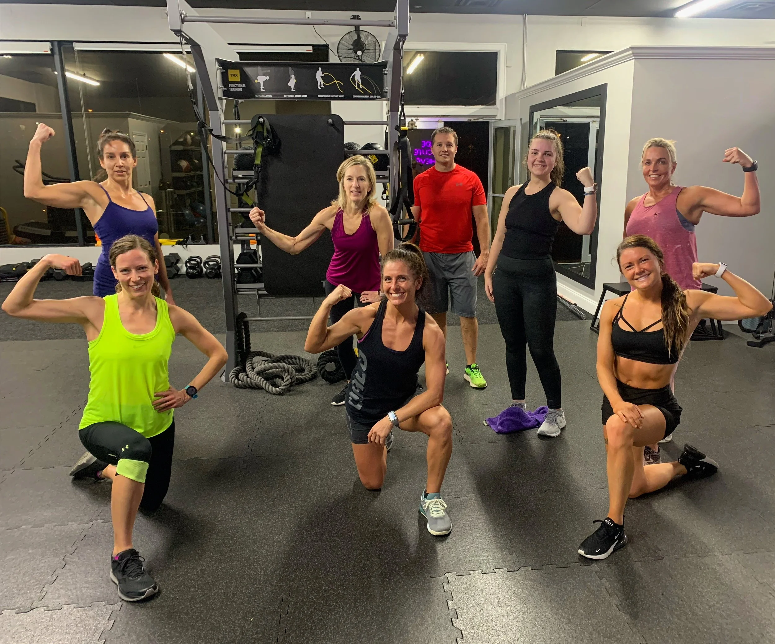 Group of nine women and one man in a gym, flexing their arm muscles, standing and kneeling on black gym mats, with fitness equipment and mirrors in the background.