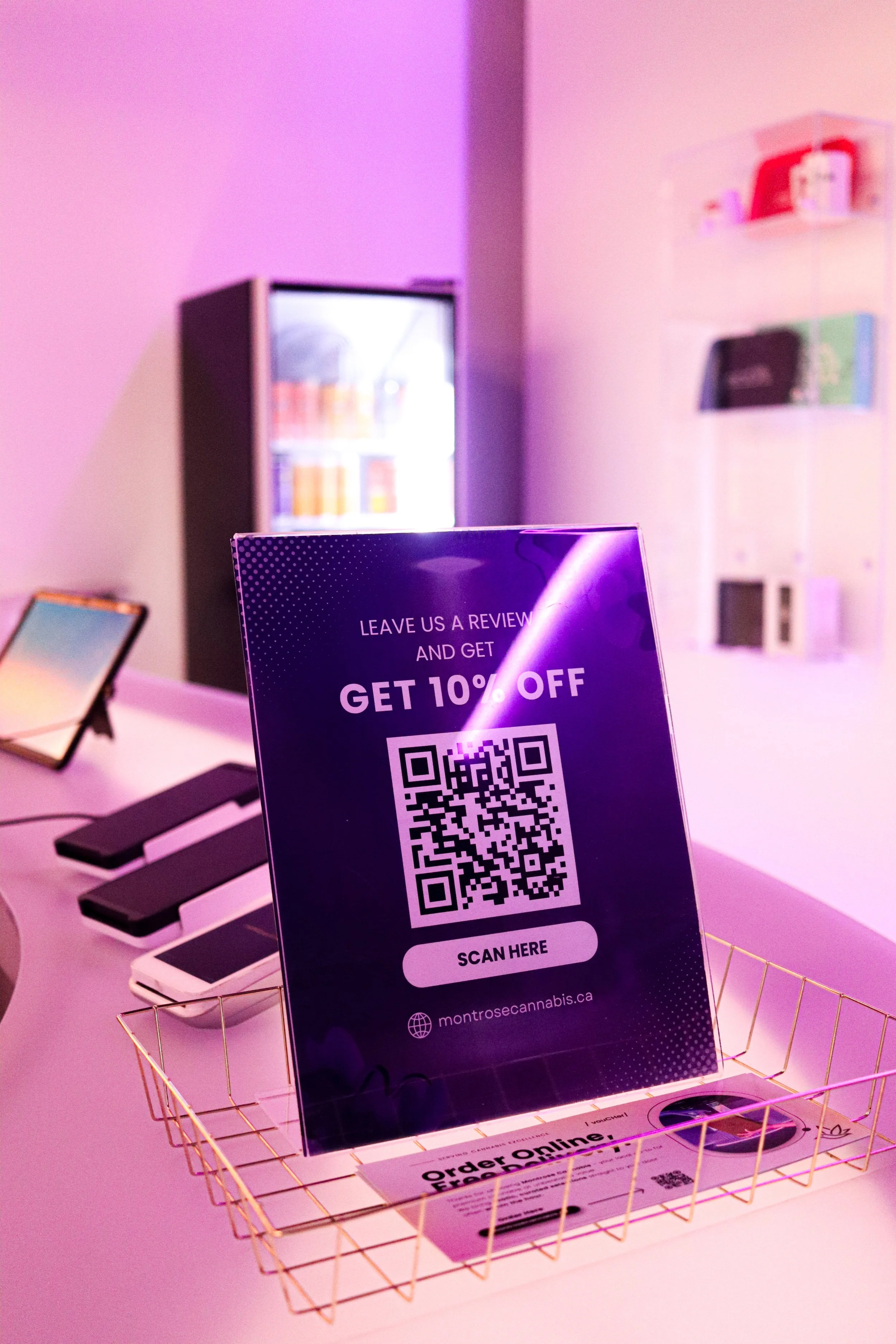 A purple sign with a QR code promoting a 10% discount for reviews, placed in a gold wire basket on a pink table. In the background, there are electronic tablets, a glass-fronted display case with beverages, and shelves with books.