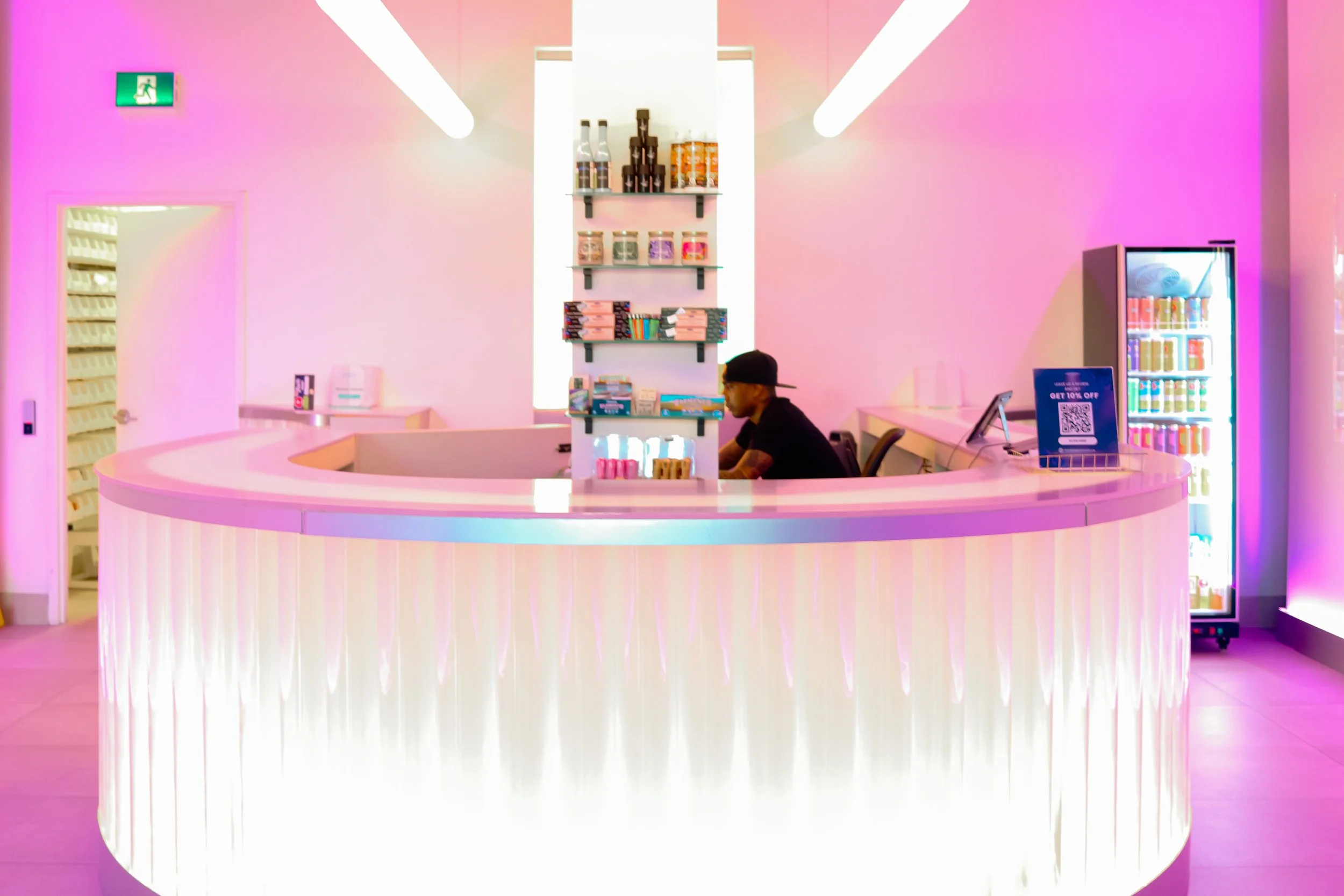 A modern, pastel-colored retail counter with a semi-circular shape, illuminated with pink LED lights. Behind the counter is a man wearing a black cap and shirt, sitting in a black chair. The background features a pink wall, a vending machine with bot