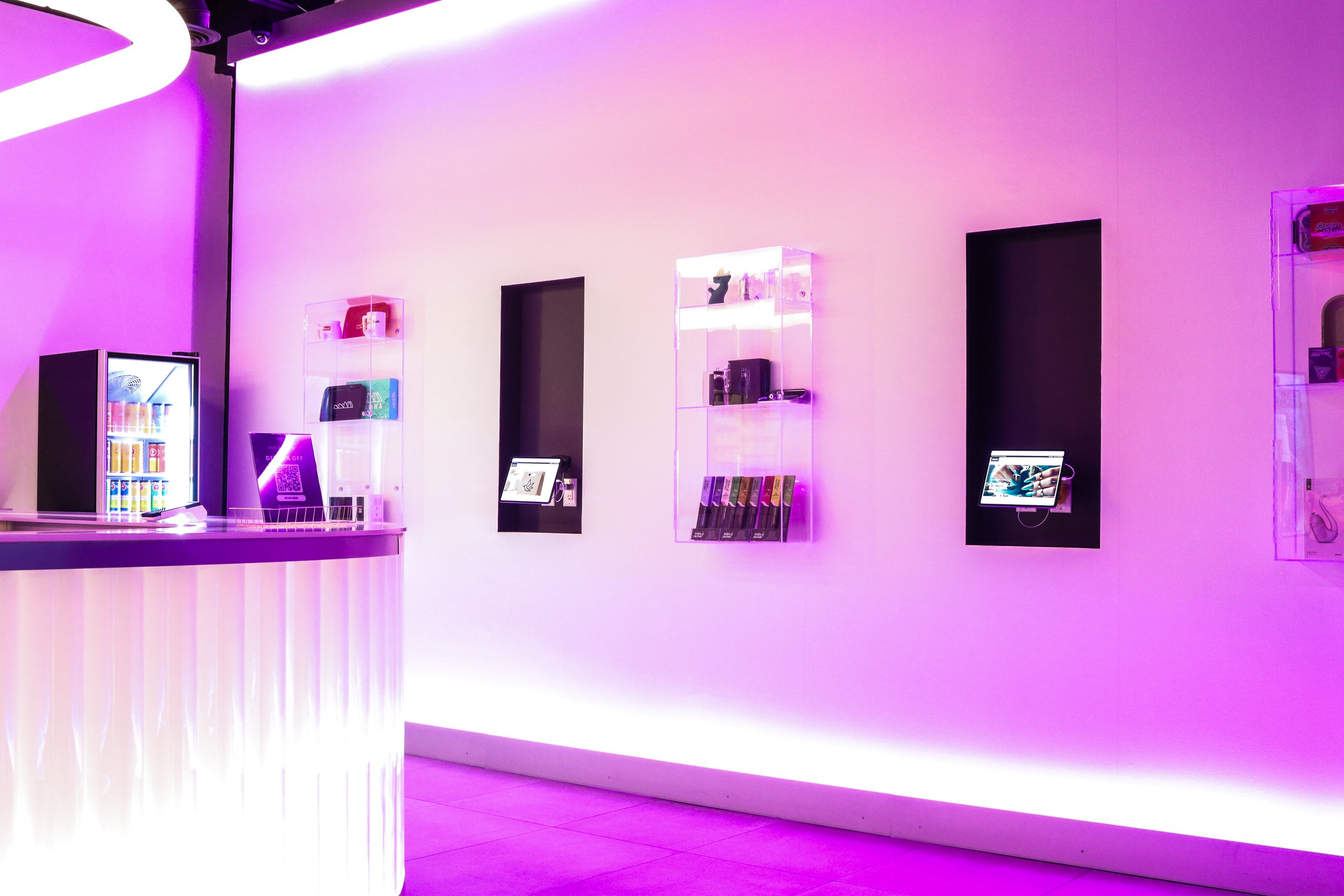 Interior of a store with purple lighting, featuring display shelves with various products, a refrigerator with beverages, and digital kiosks.