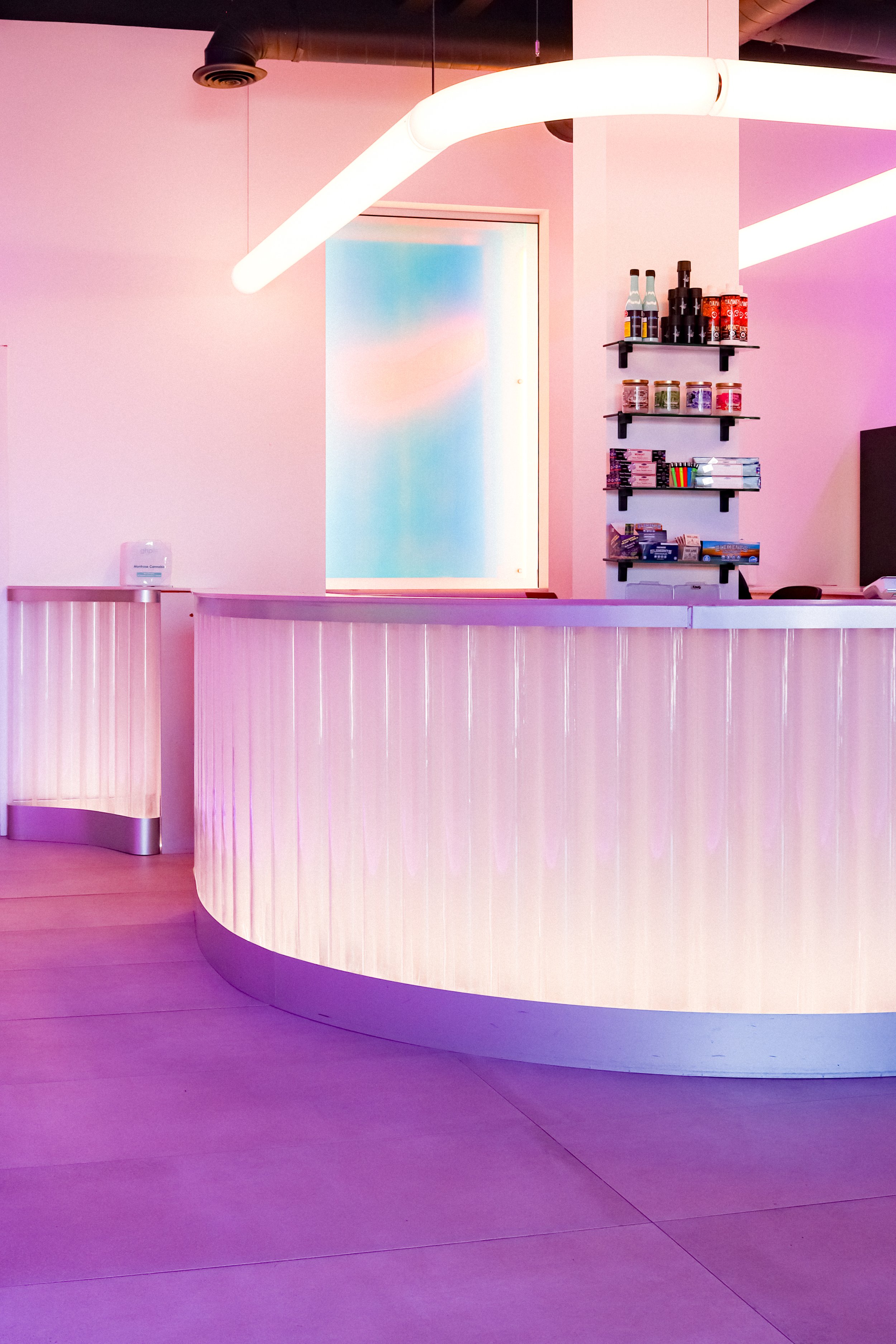 A modern, pink-themed reception counter with colorful lighting, shelves with bottles and jars, and a frosted glass window in a contemporary space.